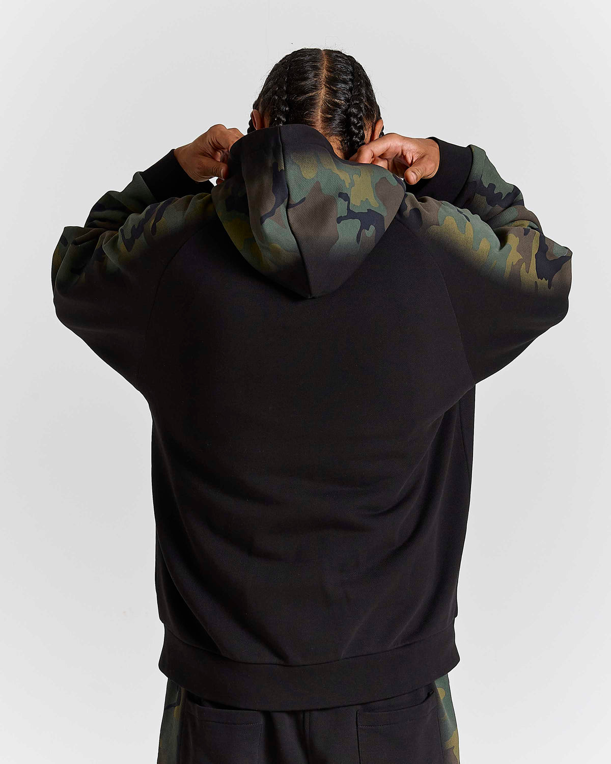 A person viewed from behind wearing a black hoodie with camouflage-patterned hood and sleeves, standing against a light gray studio background.