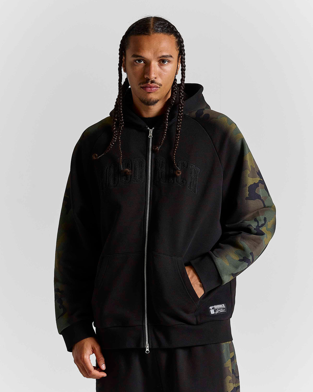 A man wearing a black zip-up hoodie with dark camouflage sleeves and a matching camouflage sweatpants, photographed against a neutral gray studio background.