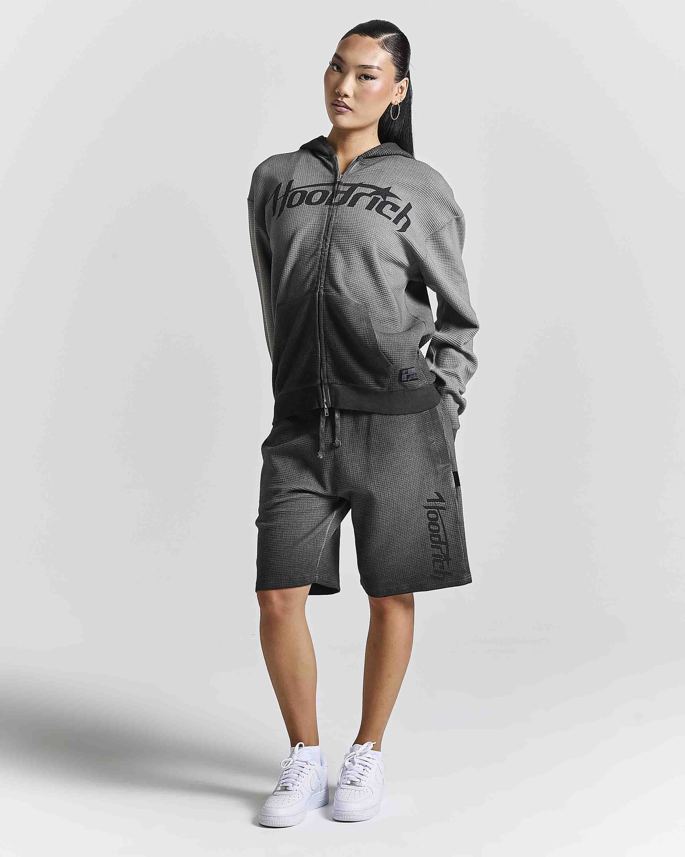A woman modeling a dark gray two-piece tracksuit featuring a zip-up hoodie with 'Hoodrich' printed across the chest and matching shorts with 'AlexRich' printed down the leg, paired with white sneakers against a light gray studio background.
