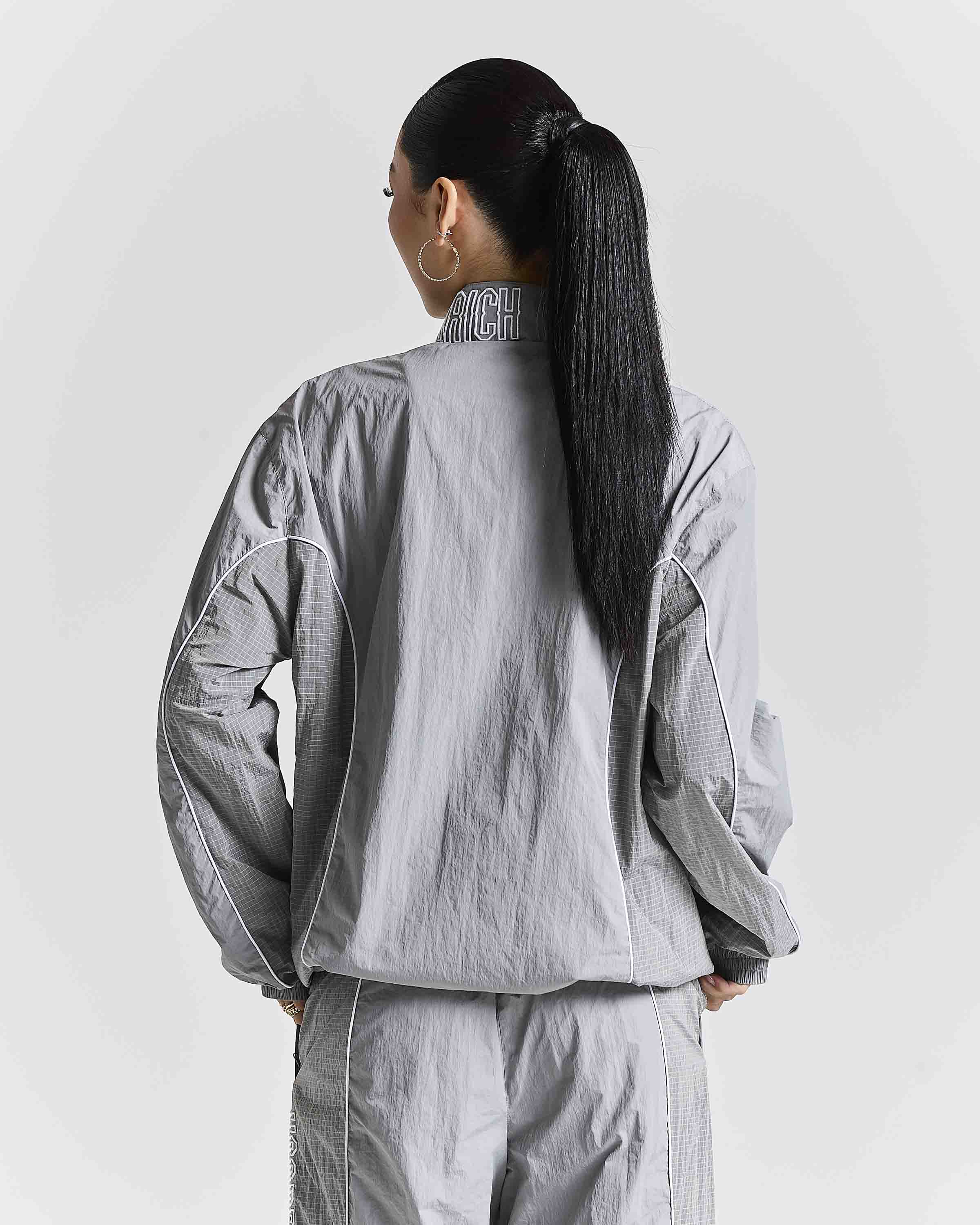 A model shown from the back wearing a light gray lightweight jacket and matching pants, featuring a high collar with 'Hoodrich' printed on it, set against a plain light gray background.