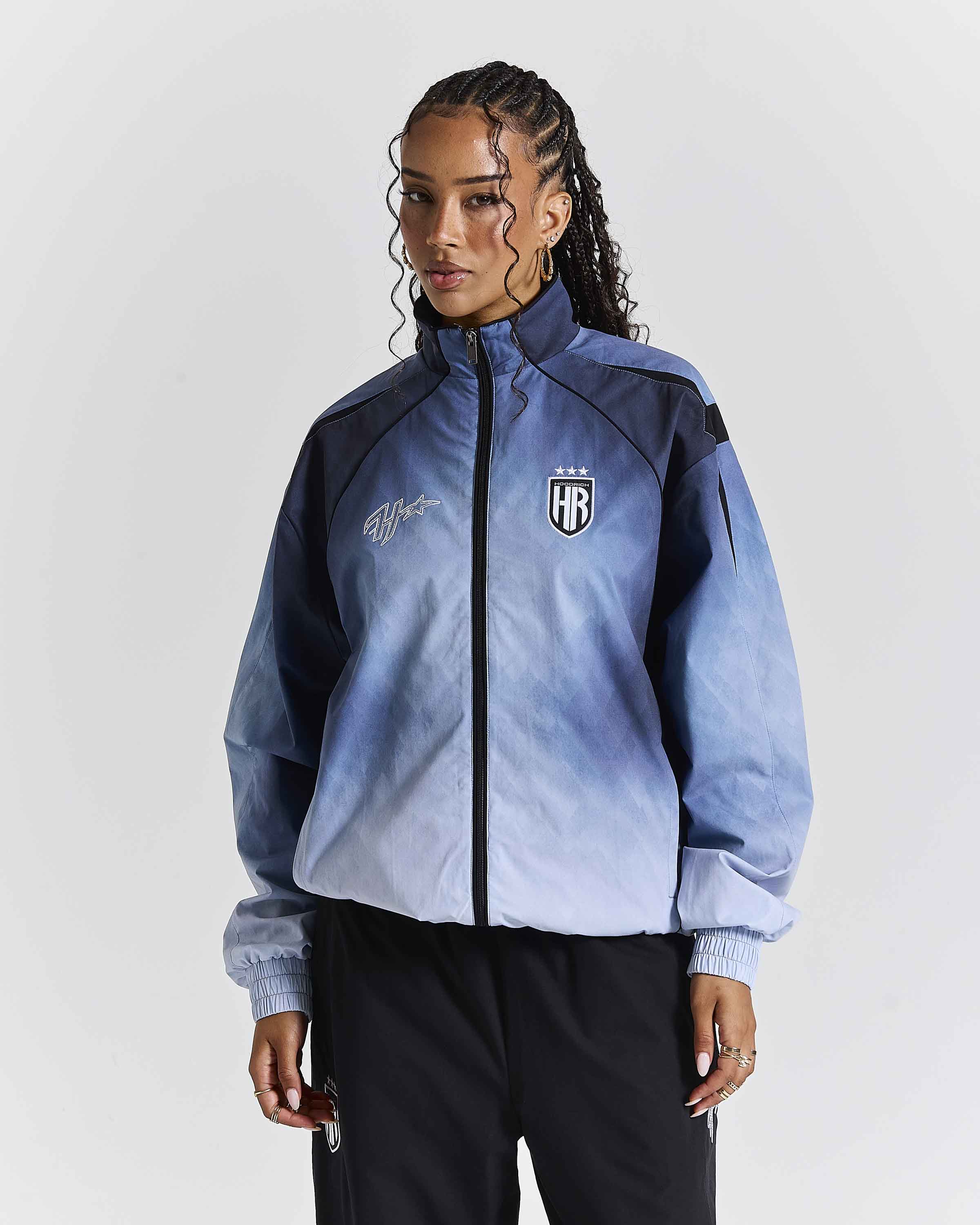 A model wearing a blue and navy gradient zip-up jacket with a black and white 'HR' shield logo on the chest, paired with black pants, against a light gray studio background.