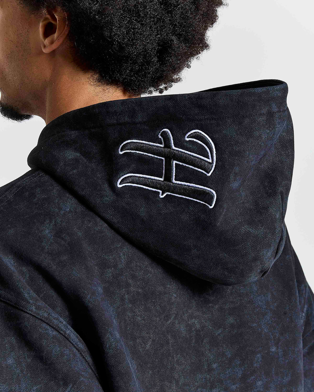 Close-up back view of a person wearing a dark hooded sweatshirt with a large white embroidered stylized symbol on the back of the hood, against a light gray background.