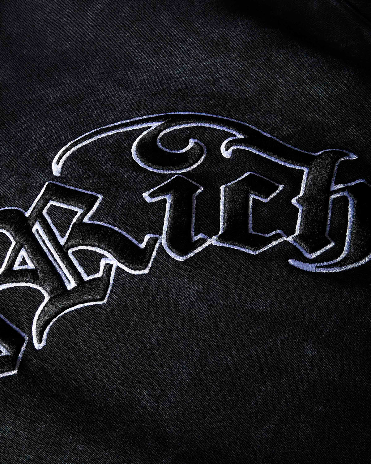 A close-up of a black textured fabric featuring a large, stylized gothic-style embroidered logo in black with a white outline.