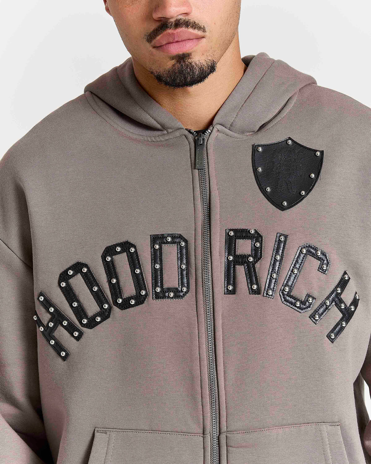 Displays a model wearing the Falcon Black 'hoodrich' branded Varso Studs Oversized Zip Through Hoodie featuring the details on the branding