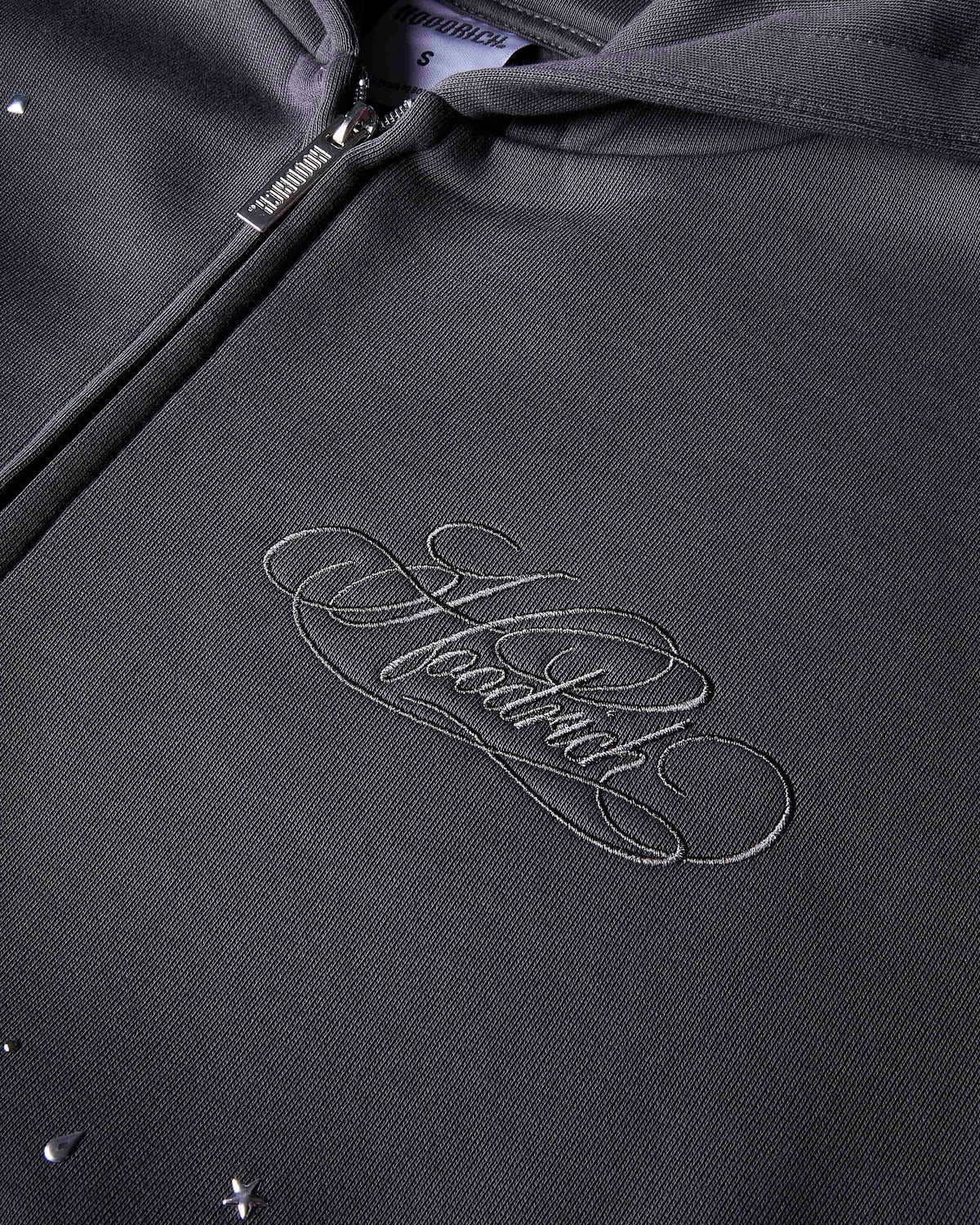 Close-up of a dark charcoal gray fabric surface with the hoodrich branding stitched scattered small reflective silver rhinestone-like studs in star, circle, and teardrop shapes.
