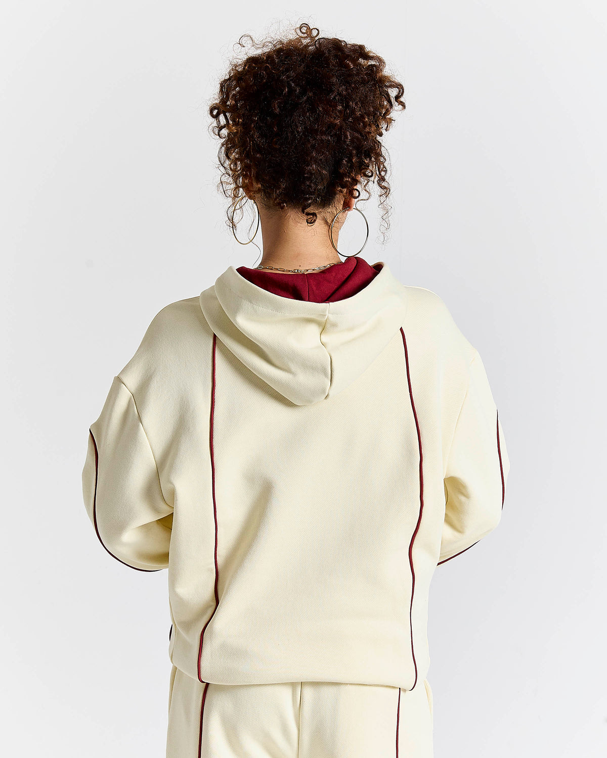 A person shown from the back wearing a cream-colored hoodie with dark red contrast piping and a dark red inner hood lining, paired with matching cream pants against a light gray studio background.
