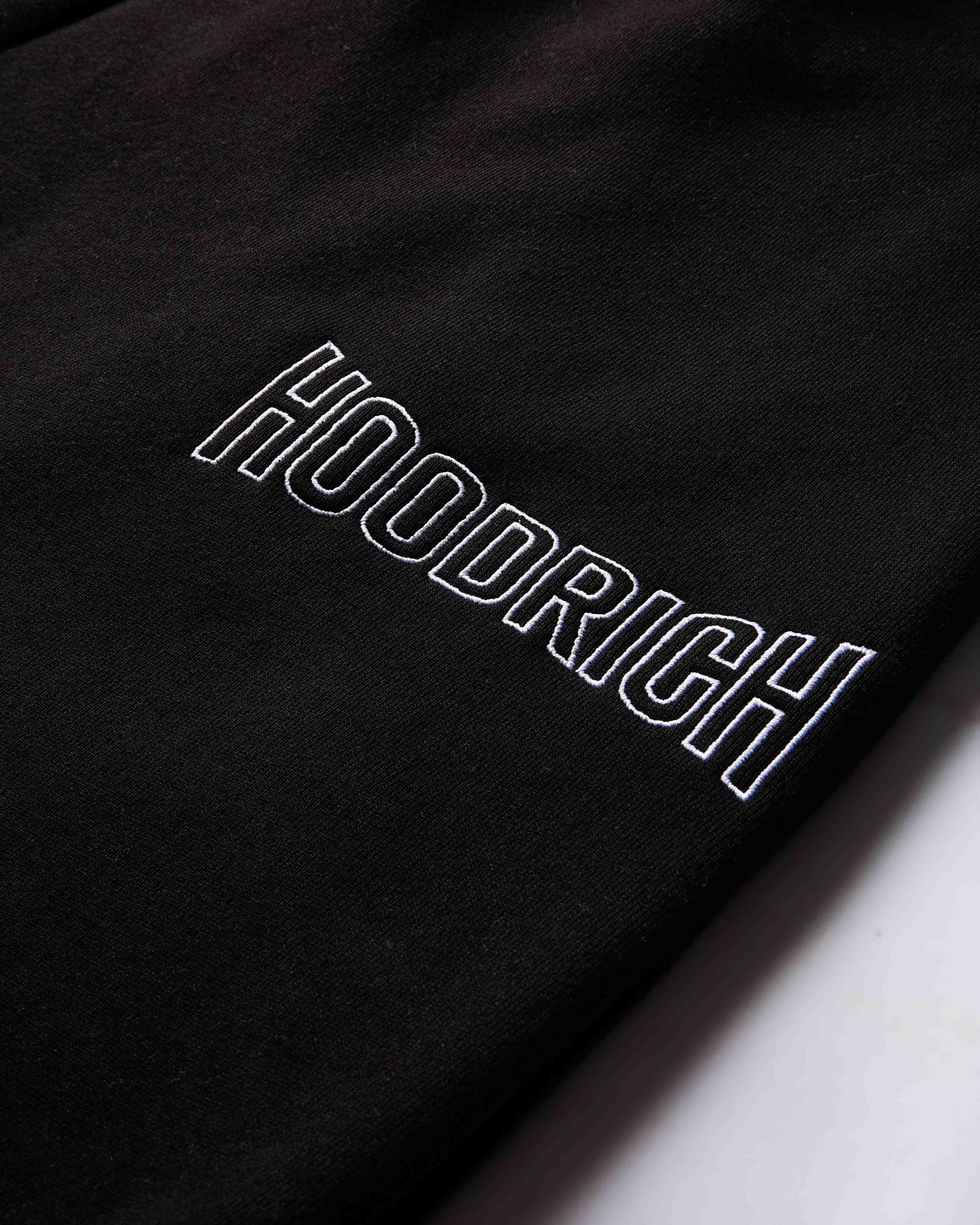 Close-up of the white outlined Hoodrich logo embroidered on the black fabric of the joggers.