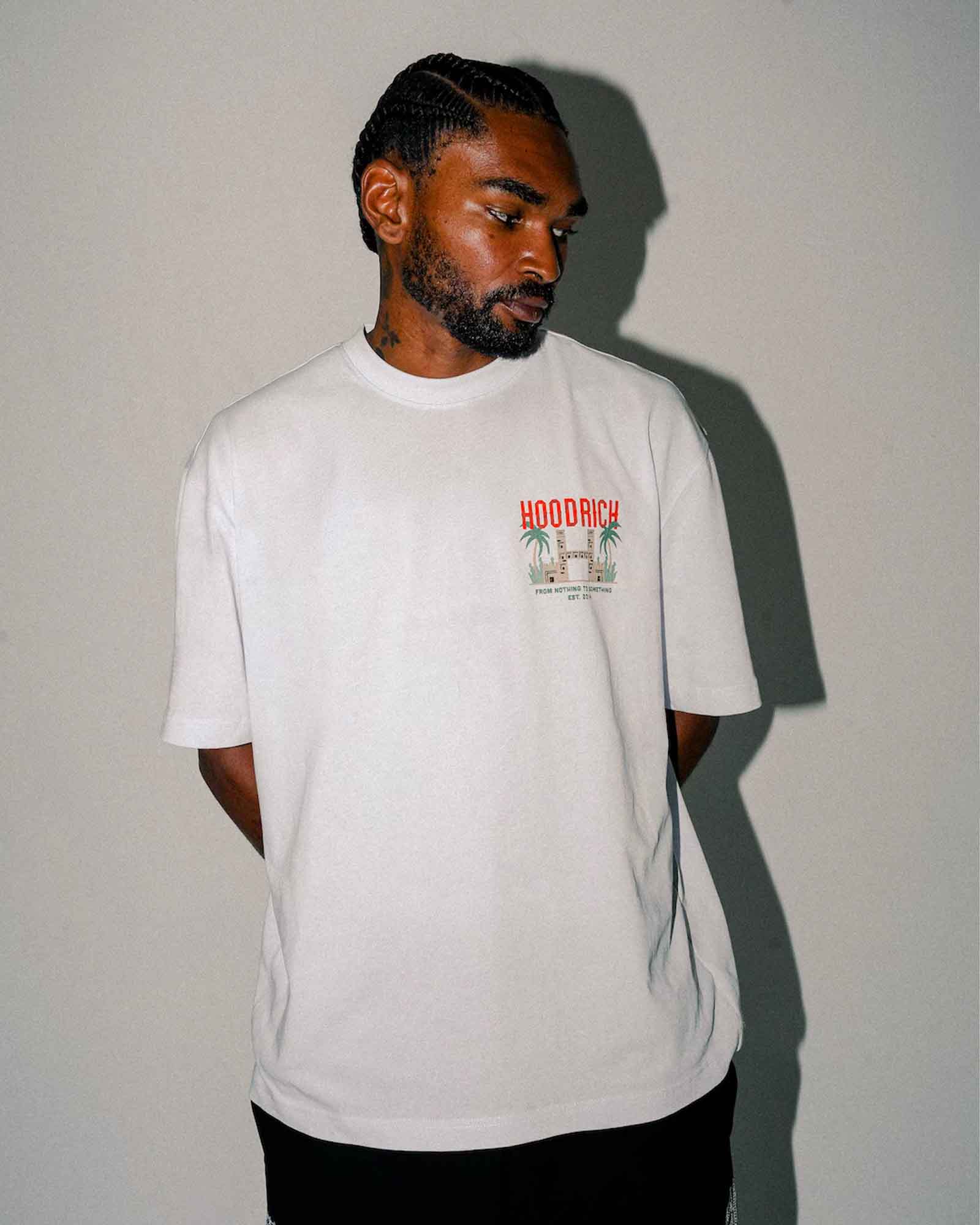 A man wearing a white crewneck t-shirt with a small red and green graphic on the left chest that reads 'HOODRICH'. The photo is taken in a dimly lit room with a strong cast shadow on the wall.