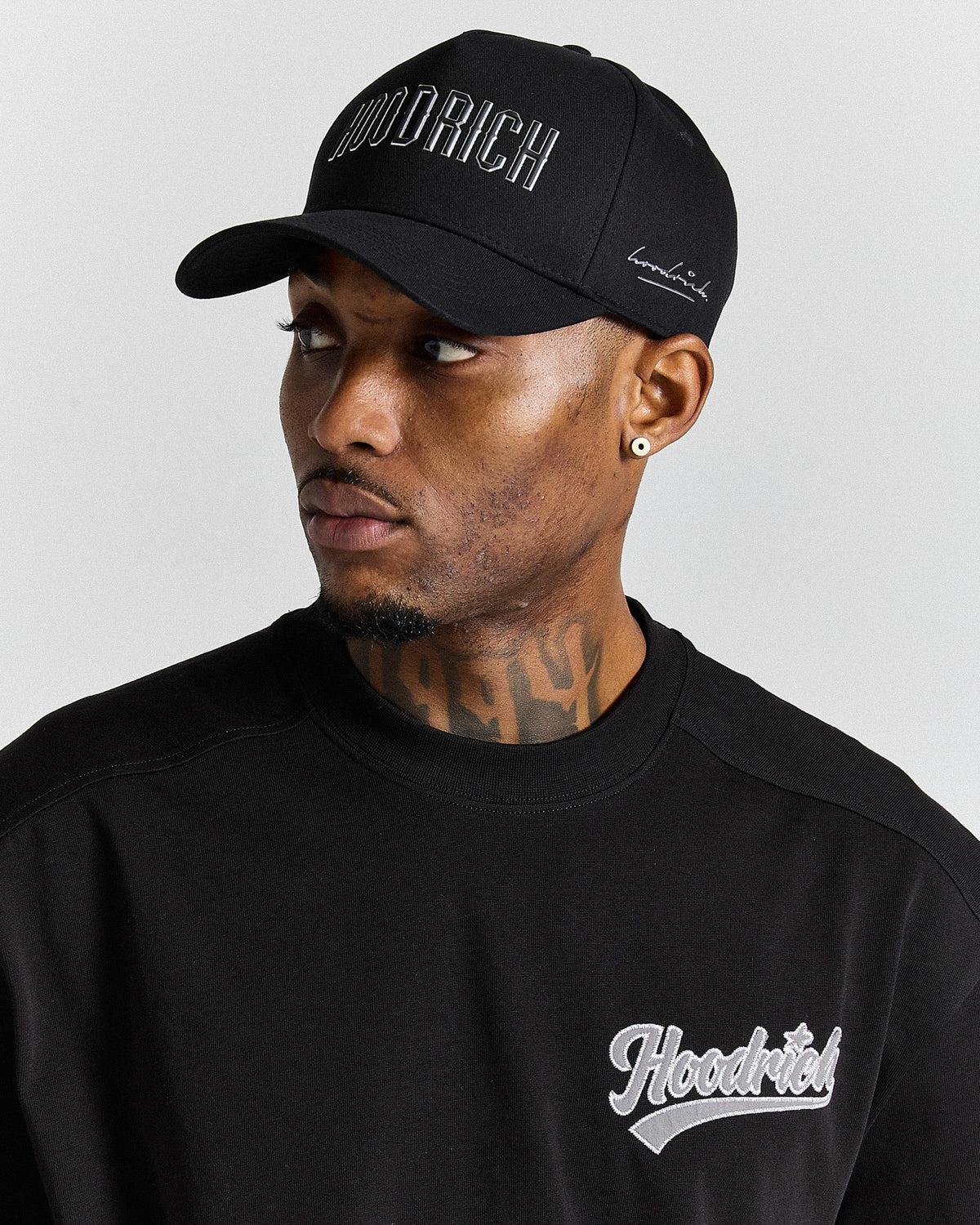 Man wearing a black 'Hoodrich' cap and sweatshirt against a gray background