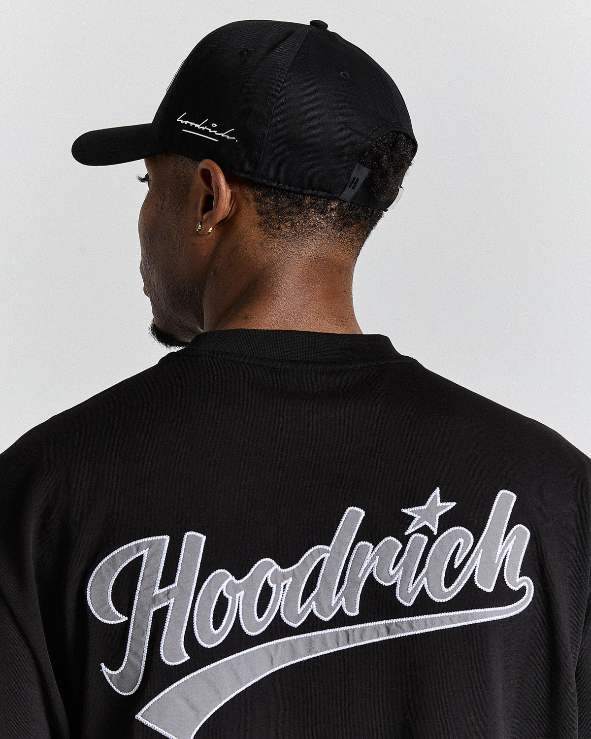 Person wearing a black cap and black 'Hoodrich' jacket on a white background