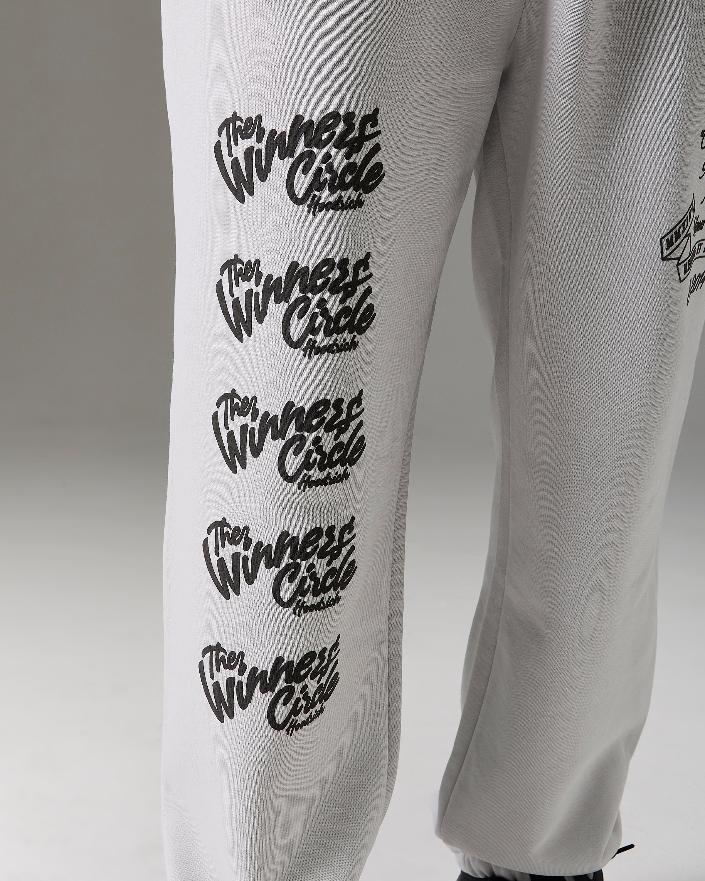 White sweatpants featuring a vertical black graphic print of the 'Winners Circle' logo repeated five times on the left leg, shown against a neutral gray background.
