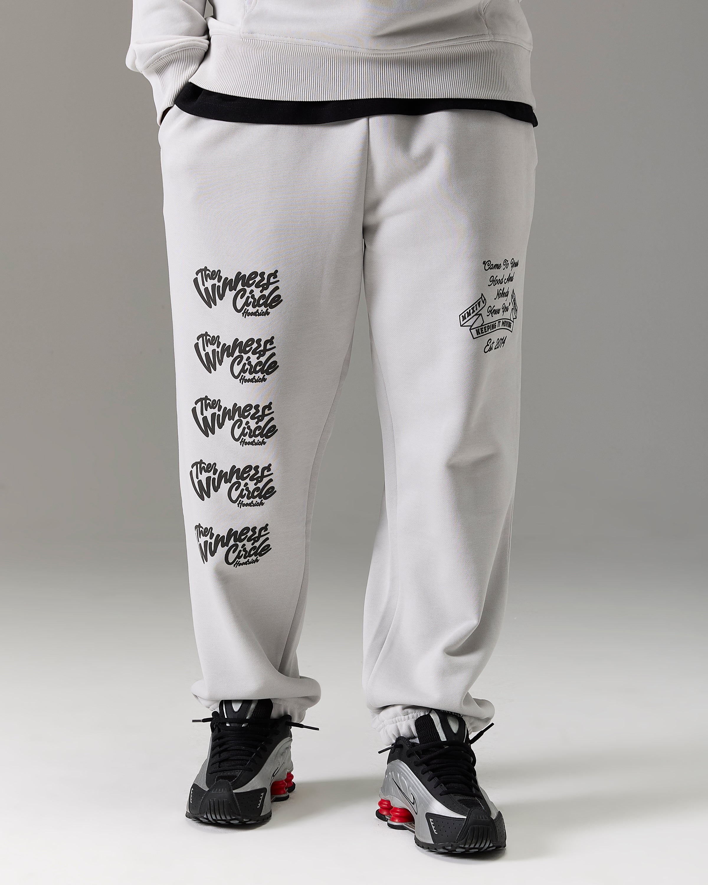 White sweatpants with black graphic prints on the legs, worn by a model with black and gray sneakers against a gray studio background.