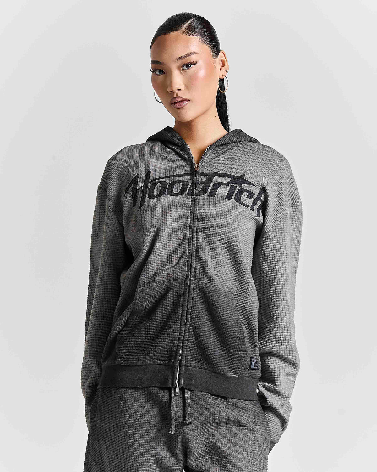 A woman modeling a dark gray waffle-knit zip-up hoodie with a black 'Hoodrich' logo across the chest, paired with matching gray pants, against a light gray studio background.