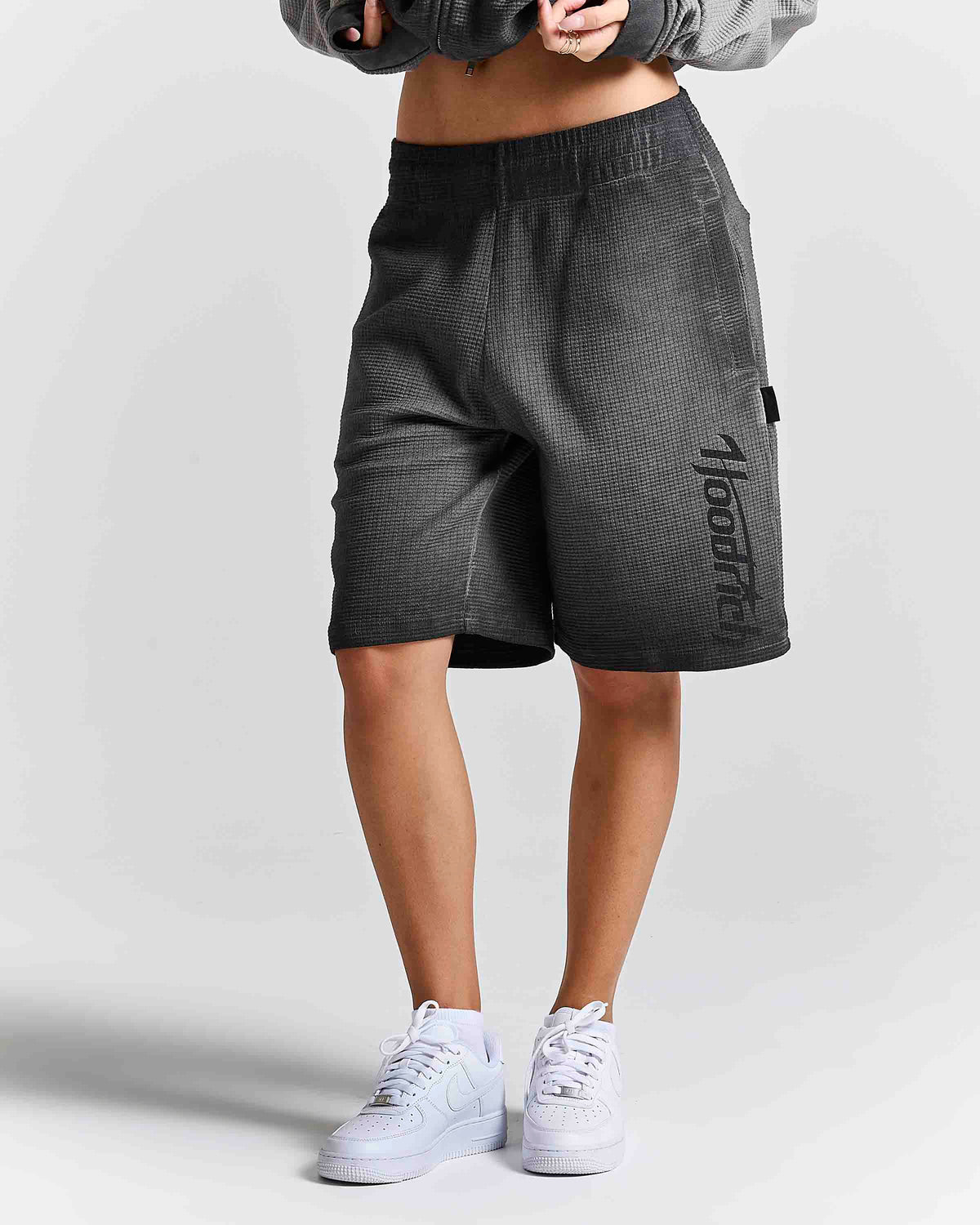 A person wearing dark gray textured athletic shorts with a vertical black 'Hoodrich' logo on the side, paired with white sneakers and a gray cropped jacket, shown from the waist down against a light gray studio background.