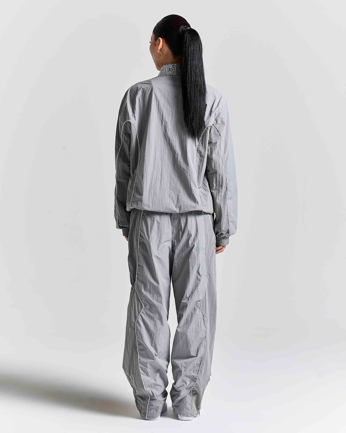 A model viewed from the back wearing a light gray matching two-piece tracksuit, consisting of a high-collar zip-up jacket and wide-leg pants, against a plain light gray studio background.