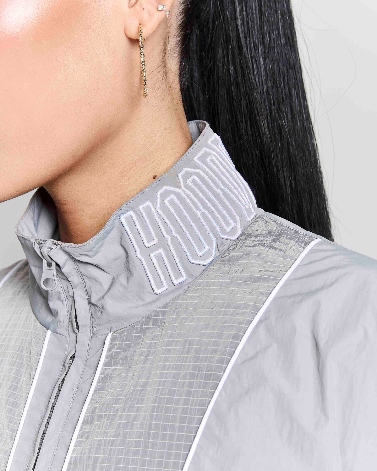 Close-up of a model wearing a light gray athletic jacket with a high collar featuring white embroidered lettering, paired with a gold dangling earring.