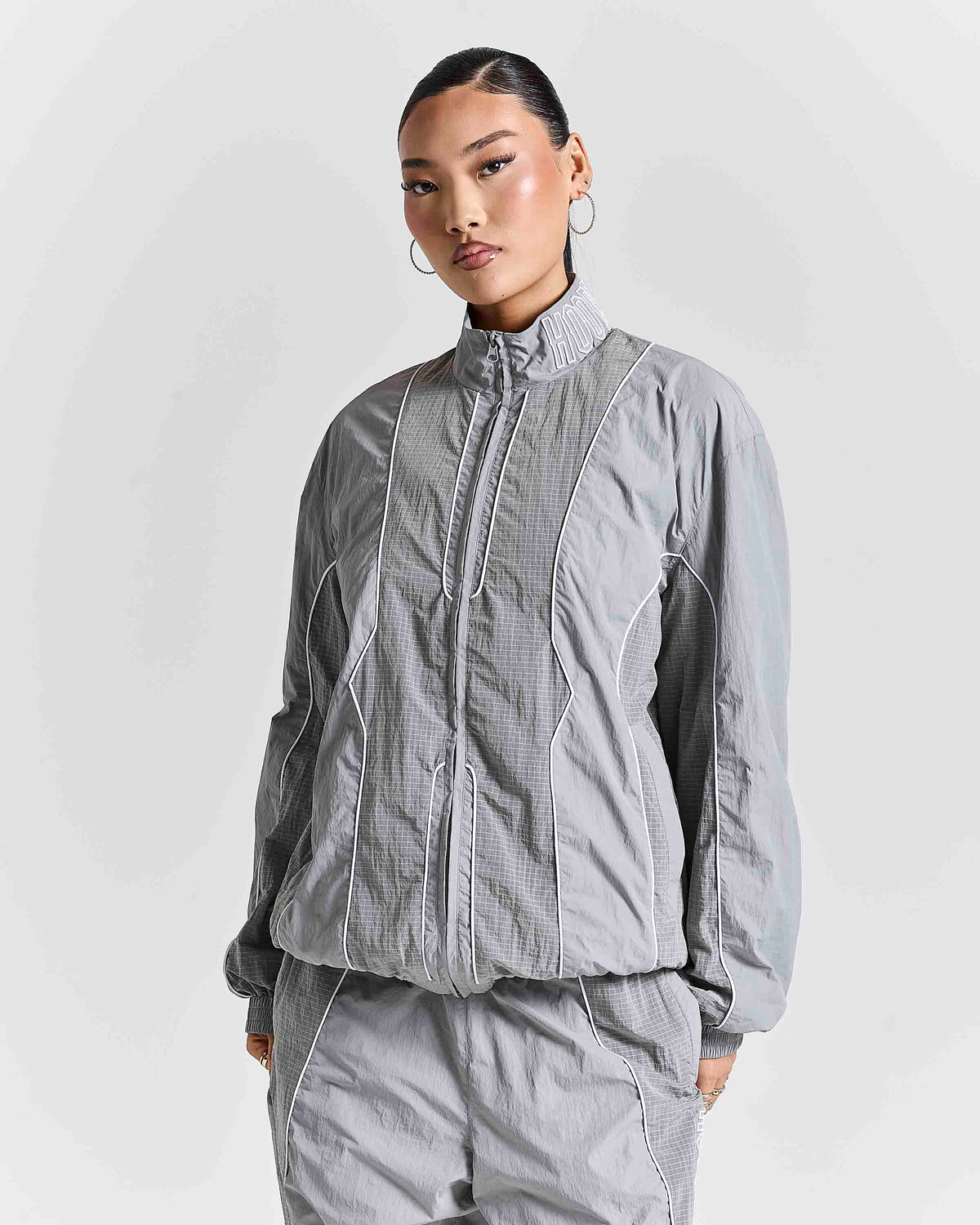 A model wearing a light gray zip-up jacket and matching gray pants against a light gray studio background.