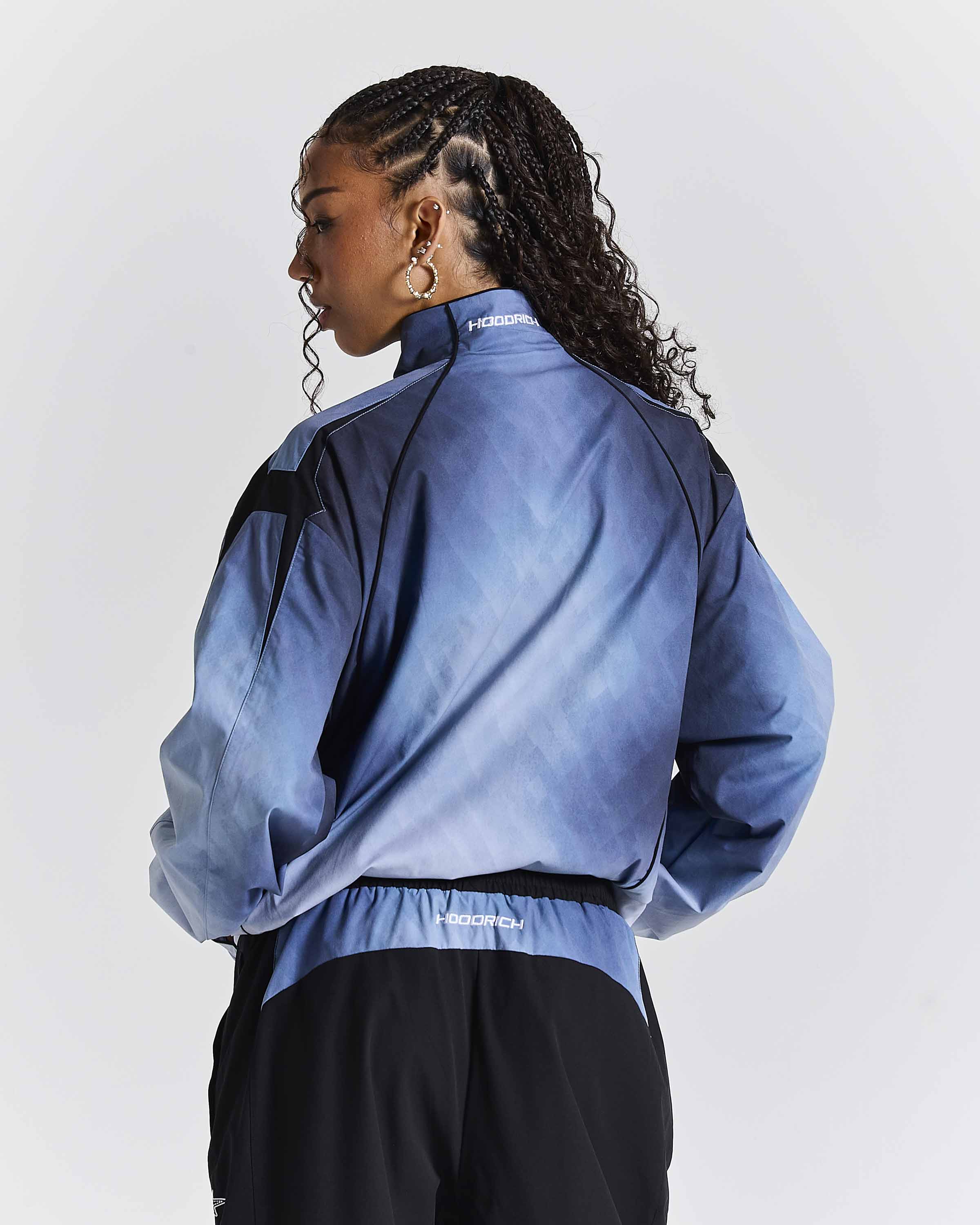 A model shown from the back wearing a blue gradient athletic jacket with black shoulder panels and black track pants against a light gray studio background.