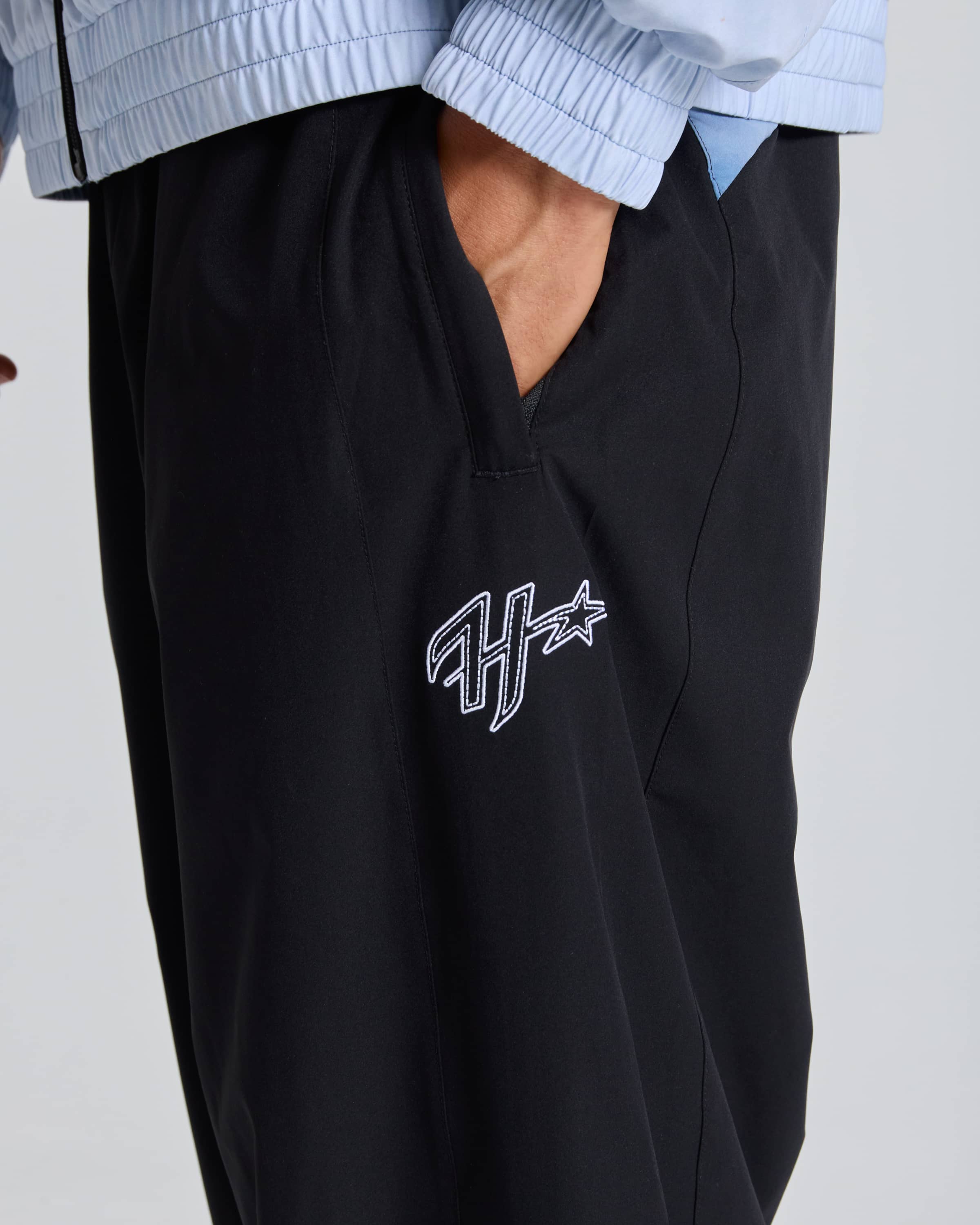 Close-up of a person wearing black jogger-style pants with a white embroidered Hoodrich logo on the left thigh, paired with a light blue collared shirt against a light gray background.