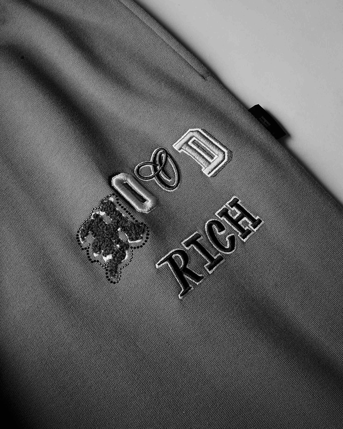 Close-up of a gray fabric surface with embroidered text reading 'HOODRICH' in metallic silver and black thread.