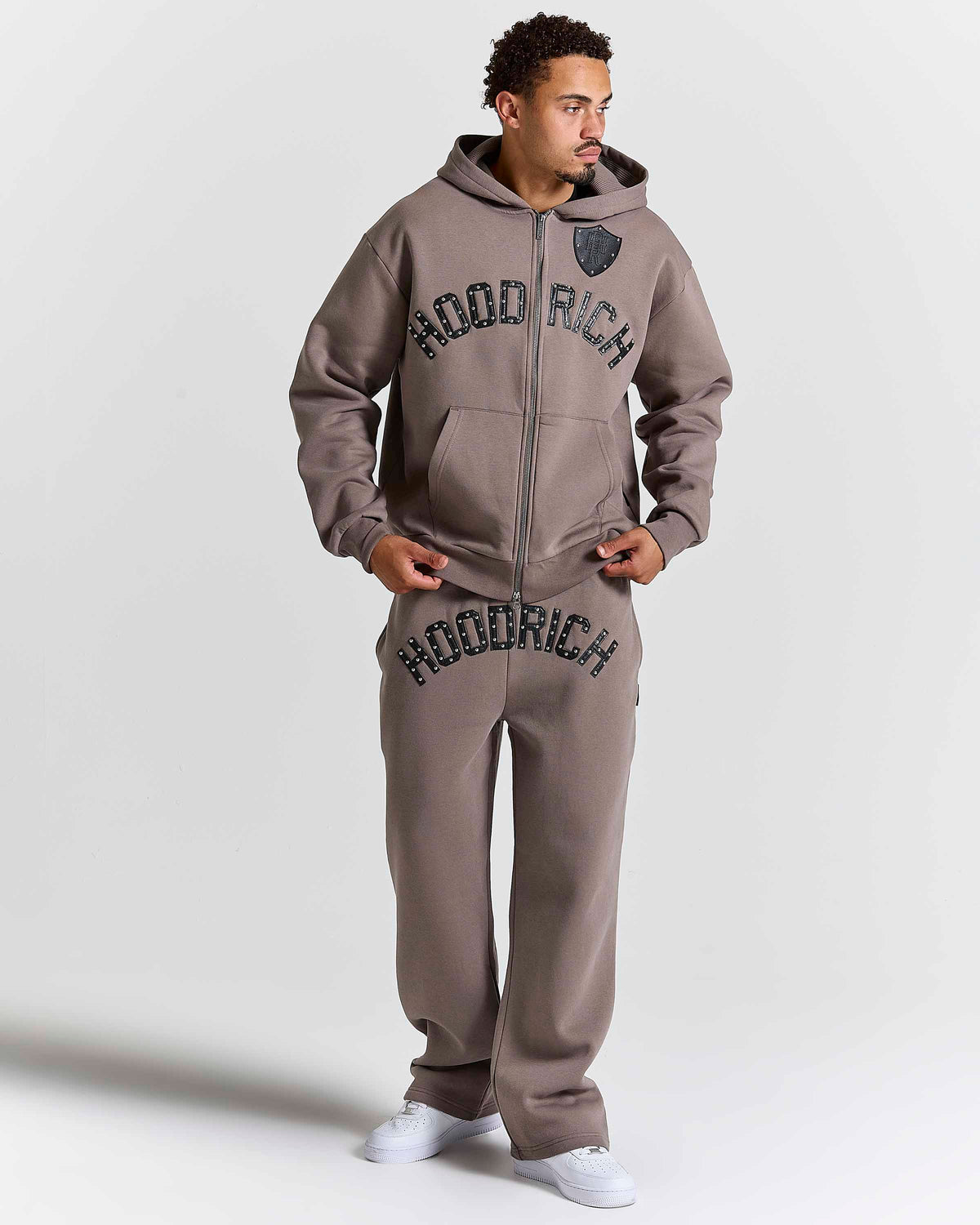 Displays a model wearing the Brown 'Hoodrich' branded men's Varso Studs Full Tracksuit 