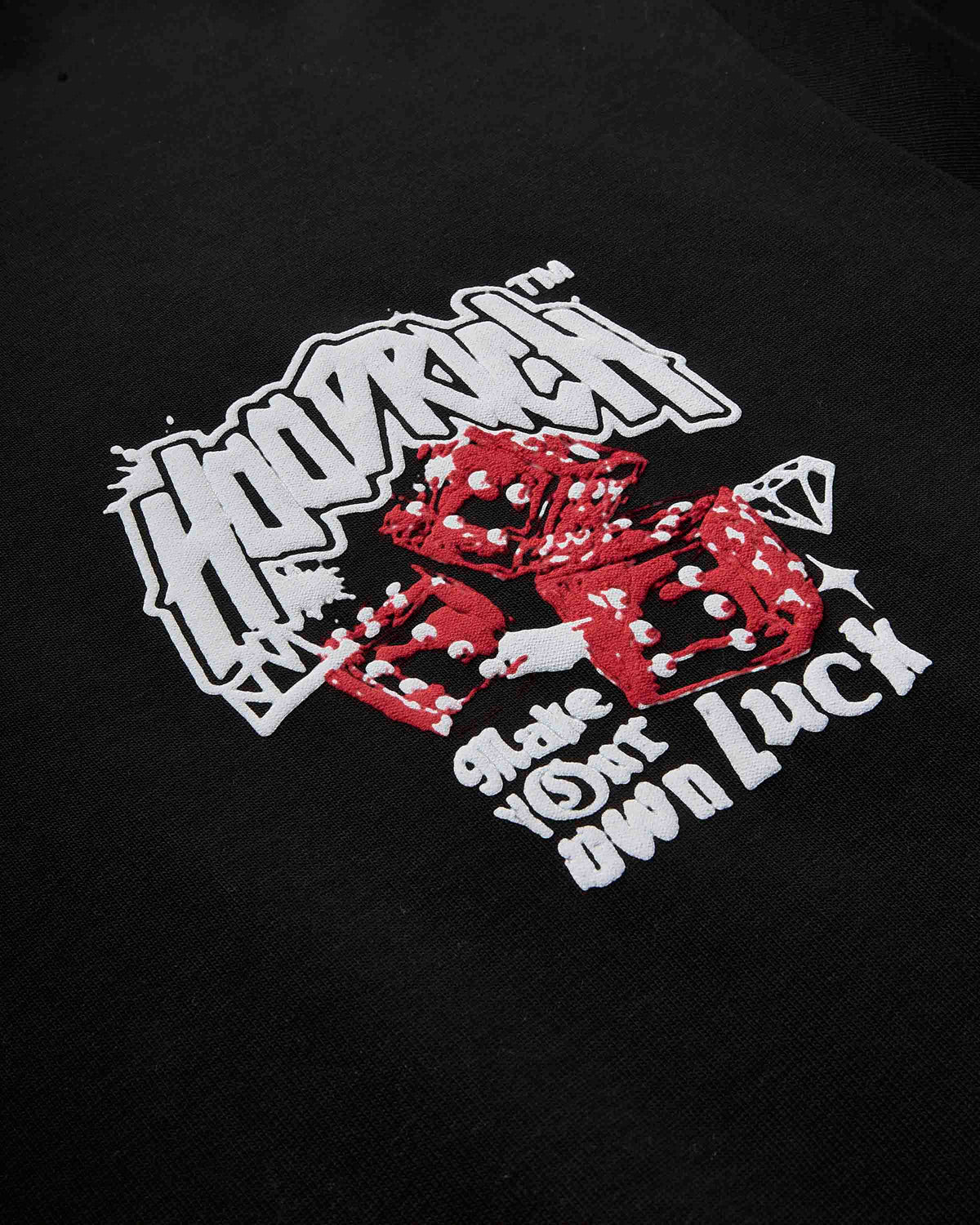 Close-up of a black t-shirt featuring a graphic print with the word 'HOODRICH' in white graffiti-style lettering, red dice, a small diamond icon, and the phrase 'Shake Your Own Luck' in white text.