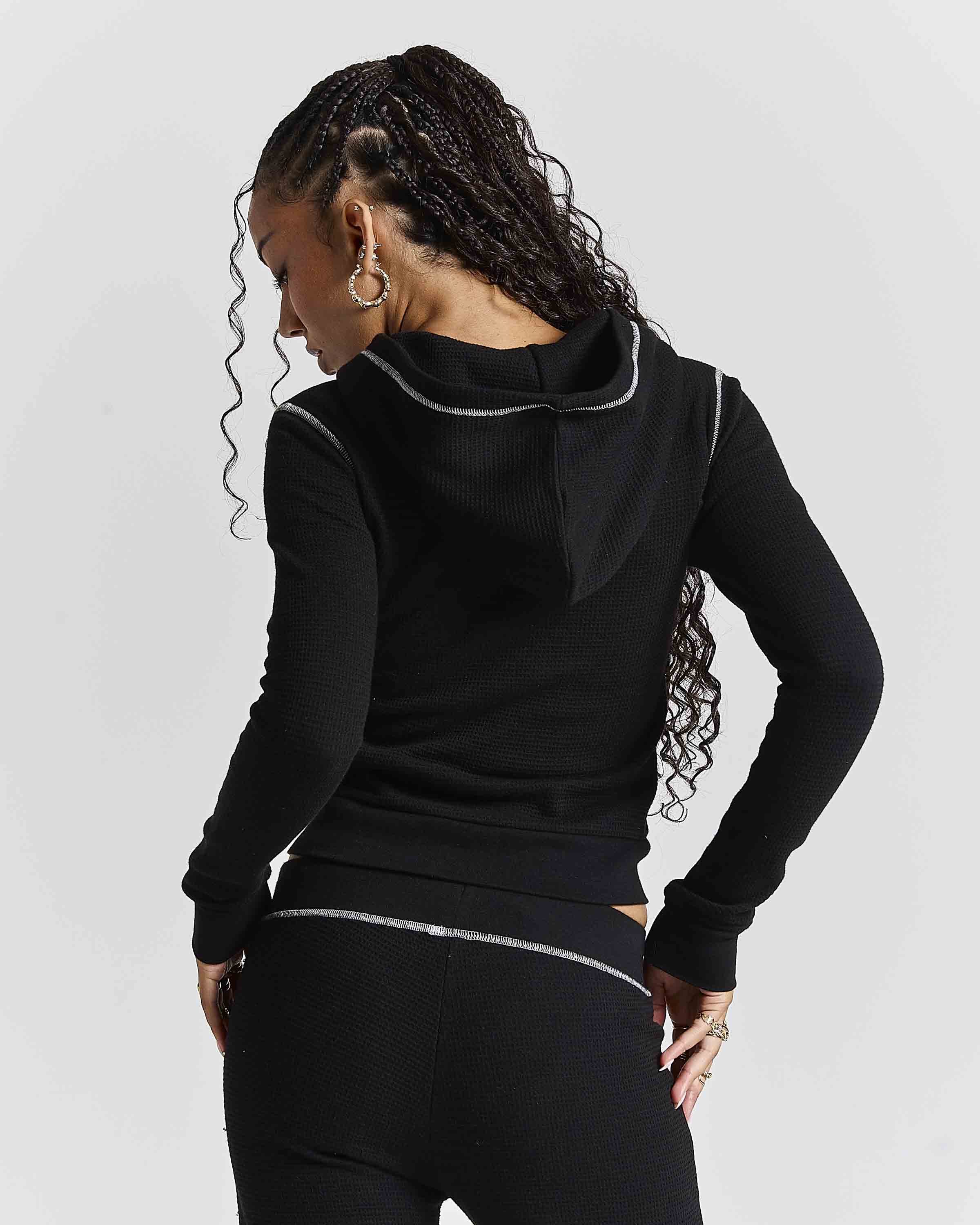 A woman shown from the back wearing a black ribbed long-sleeve hoodie with thin silver contrast piping along the hood and waist, paired with black leggings, against a light gray studio background.