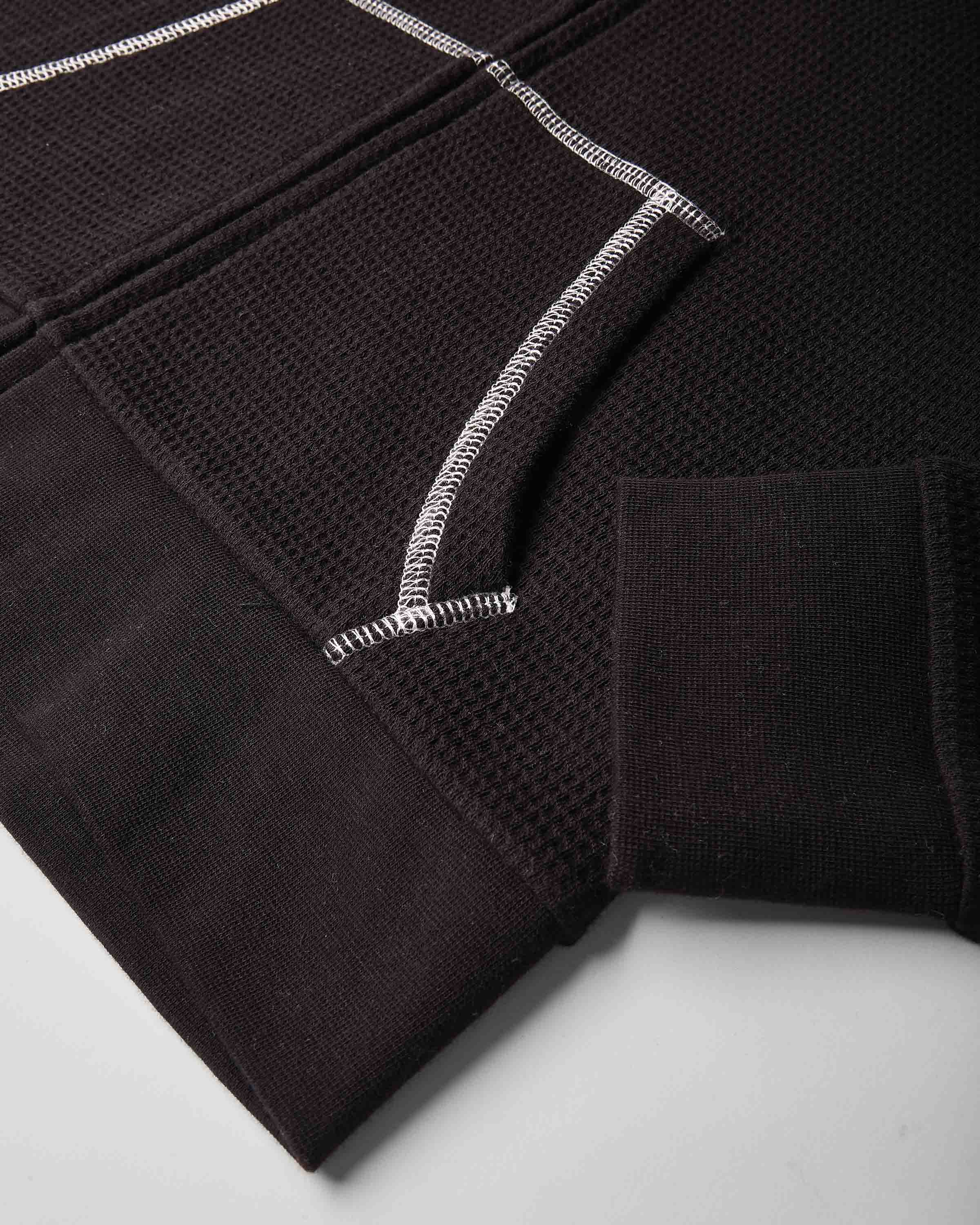 Close-up of a dark brown textured knit garment, showing a ribbed cuff, a waffle-knit panel, and contrasting white zigzag stitching on a light gray background.