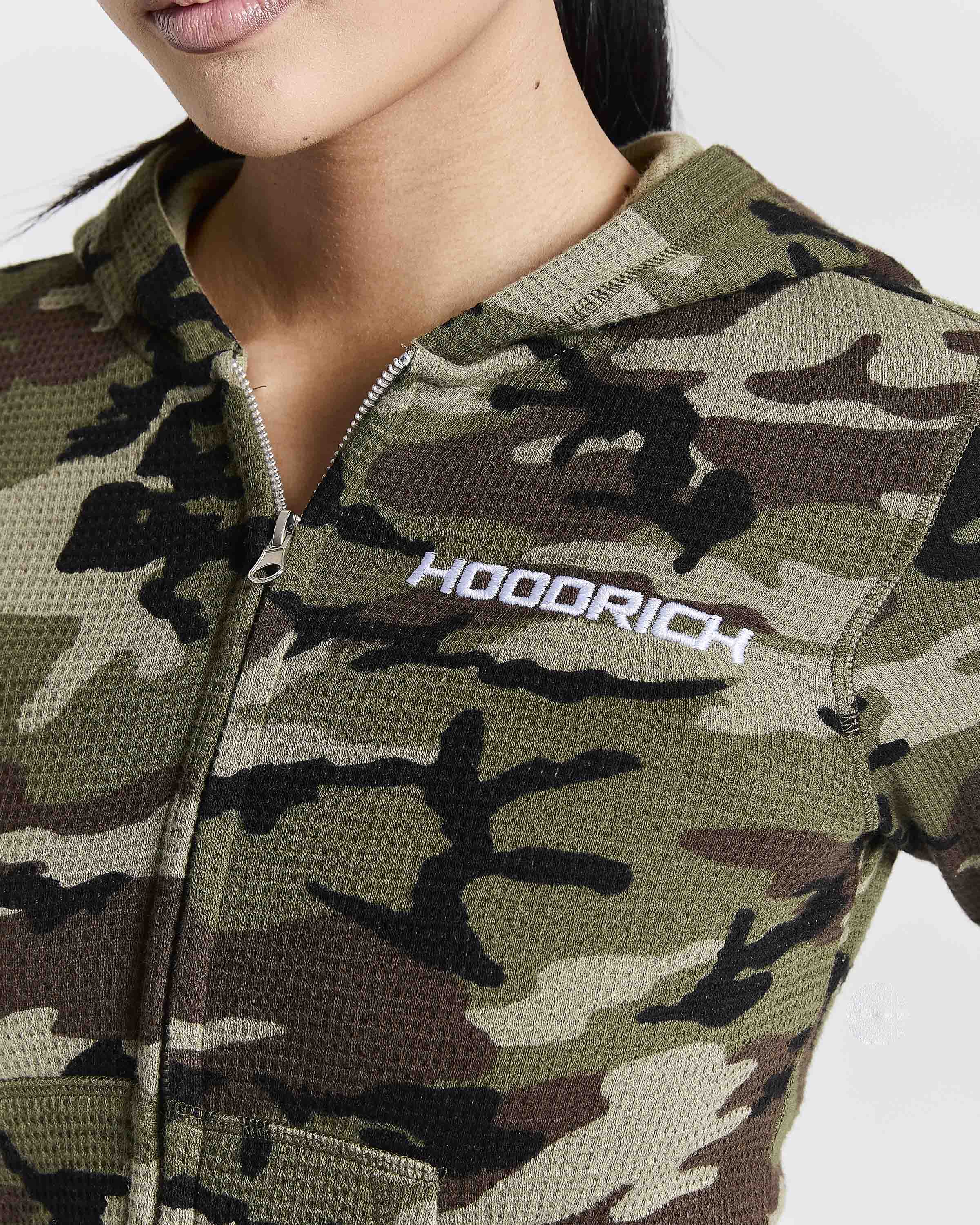 Close-up of a woman wearing a camouflage zip-up hoodie with the brand name 'HOODRICH' printed in white on the chest.