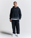 Person wearing a black hoodie and pants with 'Hoodrich' branding on a light gray background