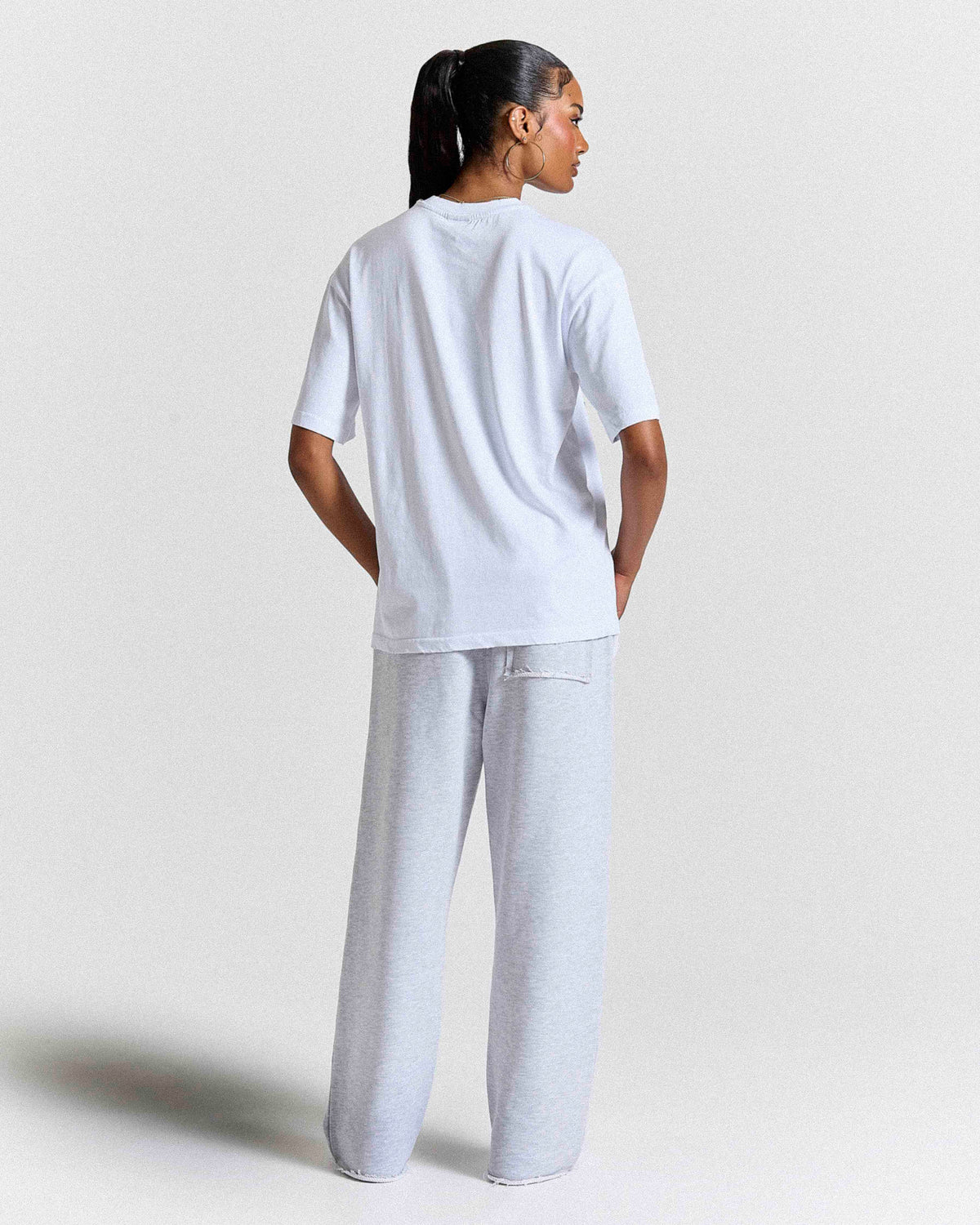 Back view of a female model wearing a white t-shirt and grey joggers on a grey studio backdrop.