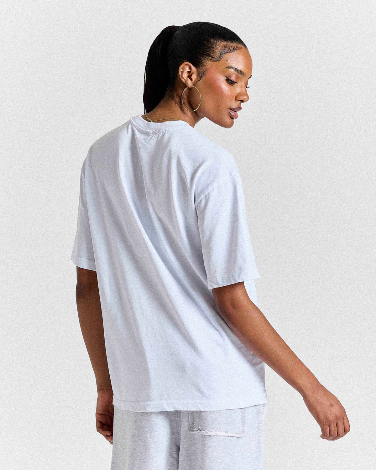 A woman shown from the back wearing an oversized white short-sleeve t-shirt and light gray sweatpants against a plain light gray studio background.