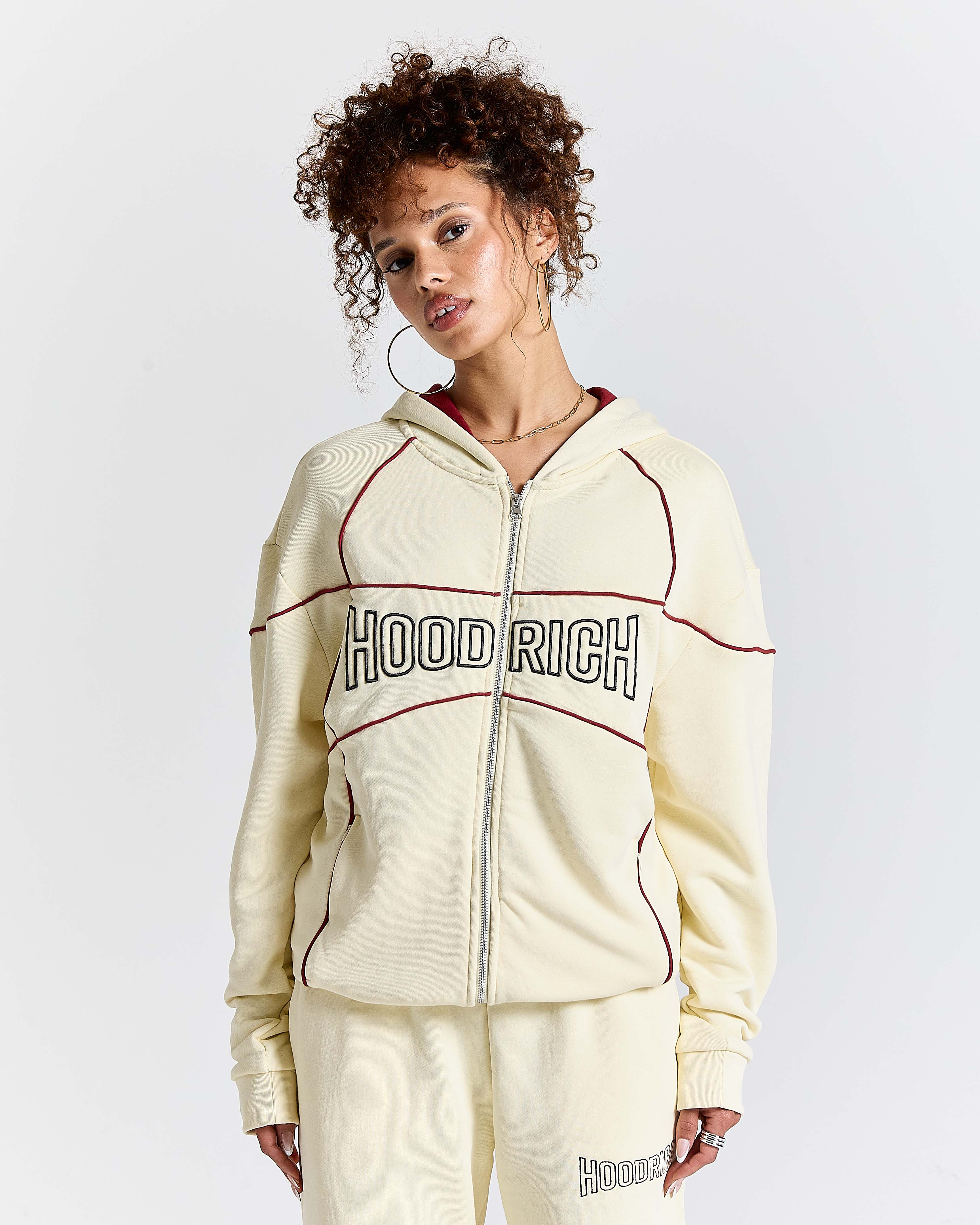 Woman wearing a cream-colored 'Hood Rich' tracksuit on a white background