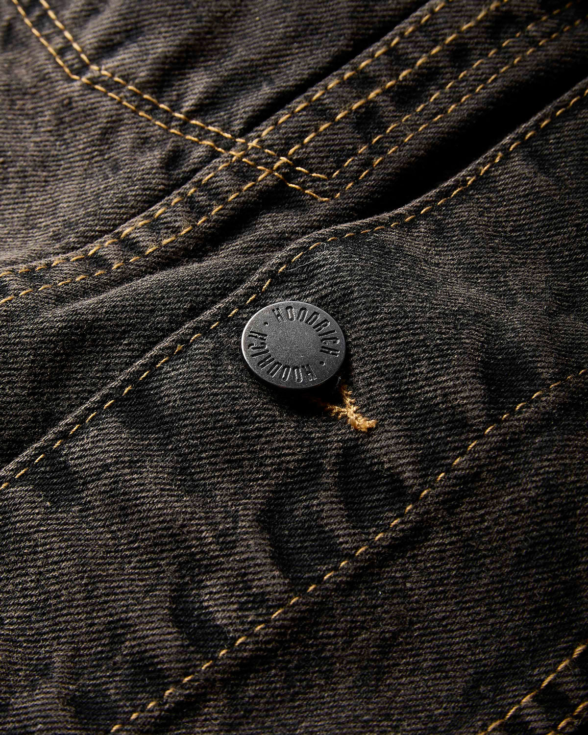 Close-up of dark charcoal gray denim fabric with contrasting tan stitching and a round black metal button embossed with 'HOODRICH'.