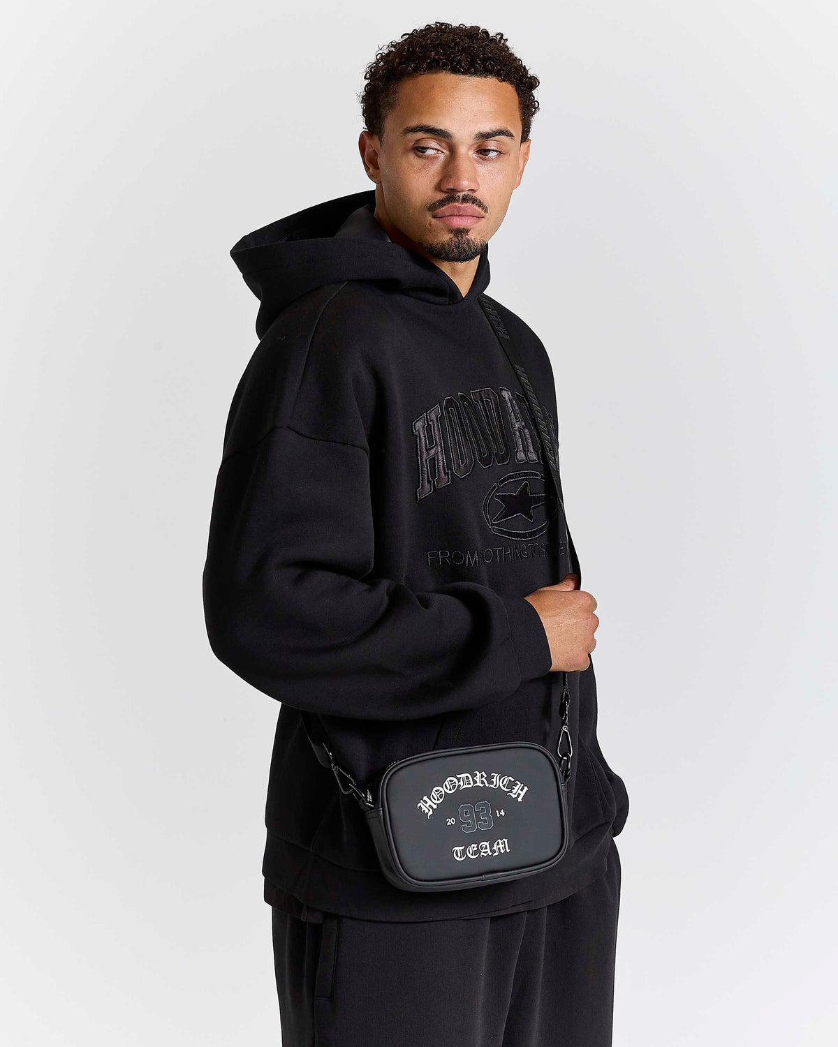 A man wearing a black hoodie and black sweatpants, carrying a black crossbody bag with 'HOODRICH TEAM' printed on it, against a light gray studio background.