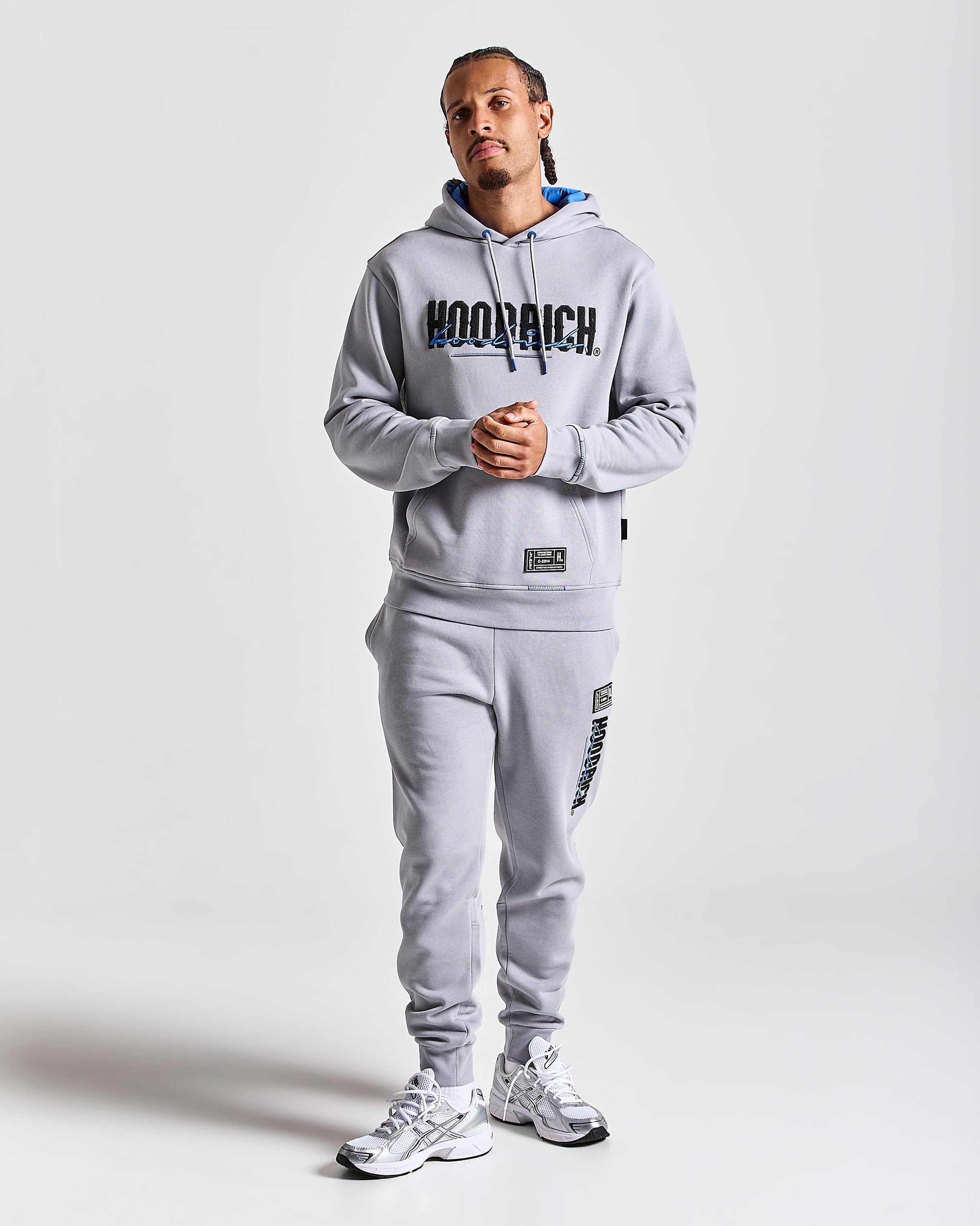 A man modeling a light gray Hoodrich hoodie and matching sweatpants set, featuring a large black and blue 'HOODRICH' logo across the chest and smaller branding on the pants, against a light gray studio background.