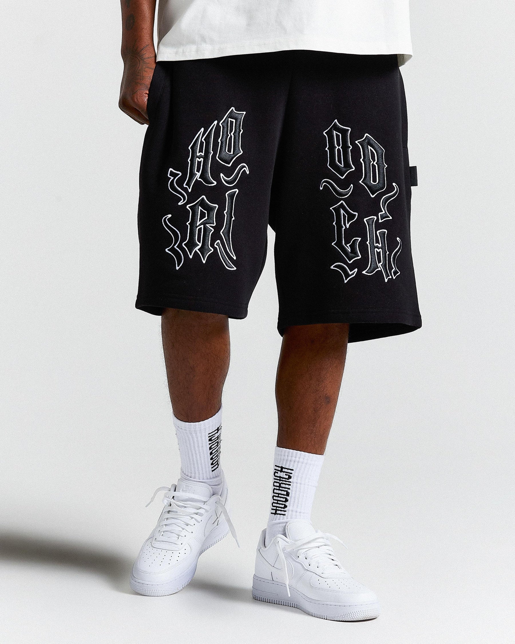 Person wearing black shorts with white text and white sneakers on a light gray background