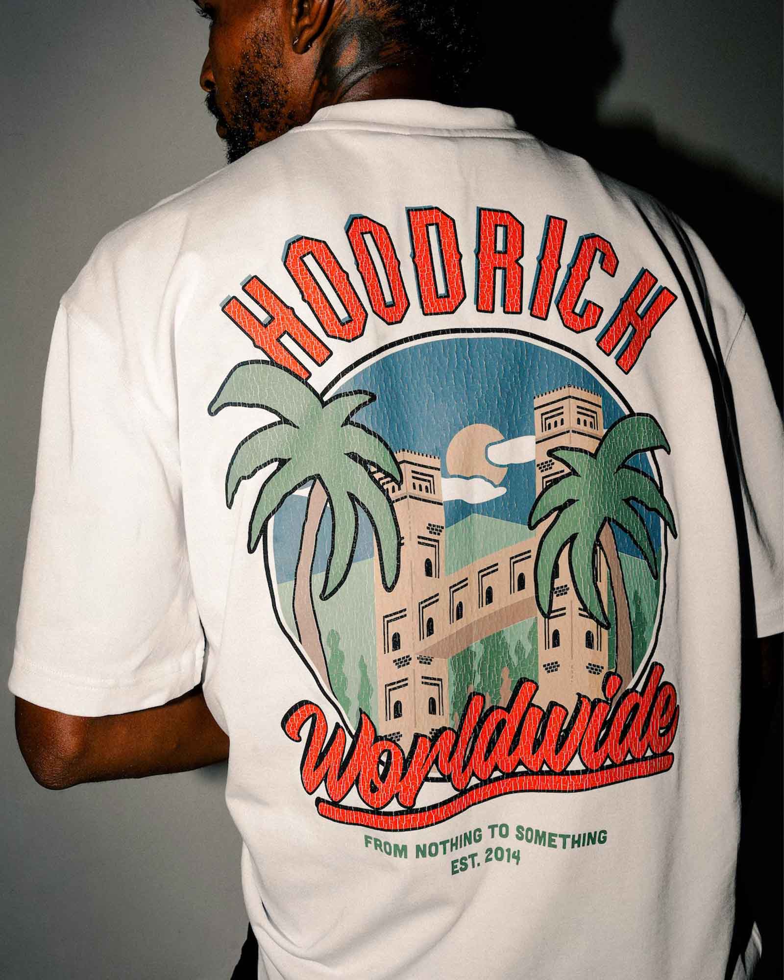 A man viewed from behind wearing a white t-shirt with a large graphic print on the back. The graphic features the text 'HOODRICH Worldwide' in red, with a central illustration of a building, two palm trees, and a blue sky background.