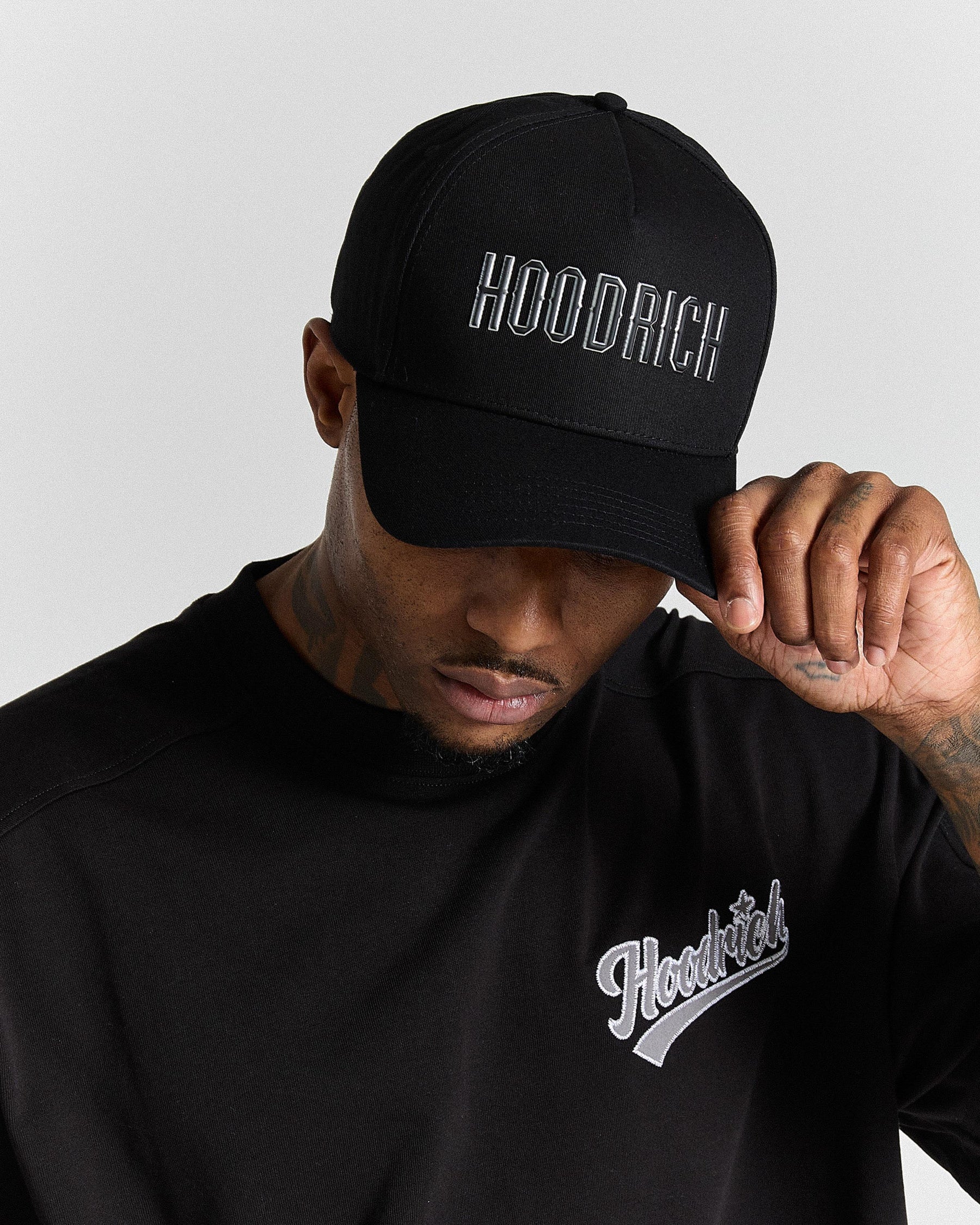 Person wearing a black cap and black hoodie with 'Hoodrich' branding on a light gray background