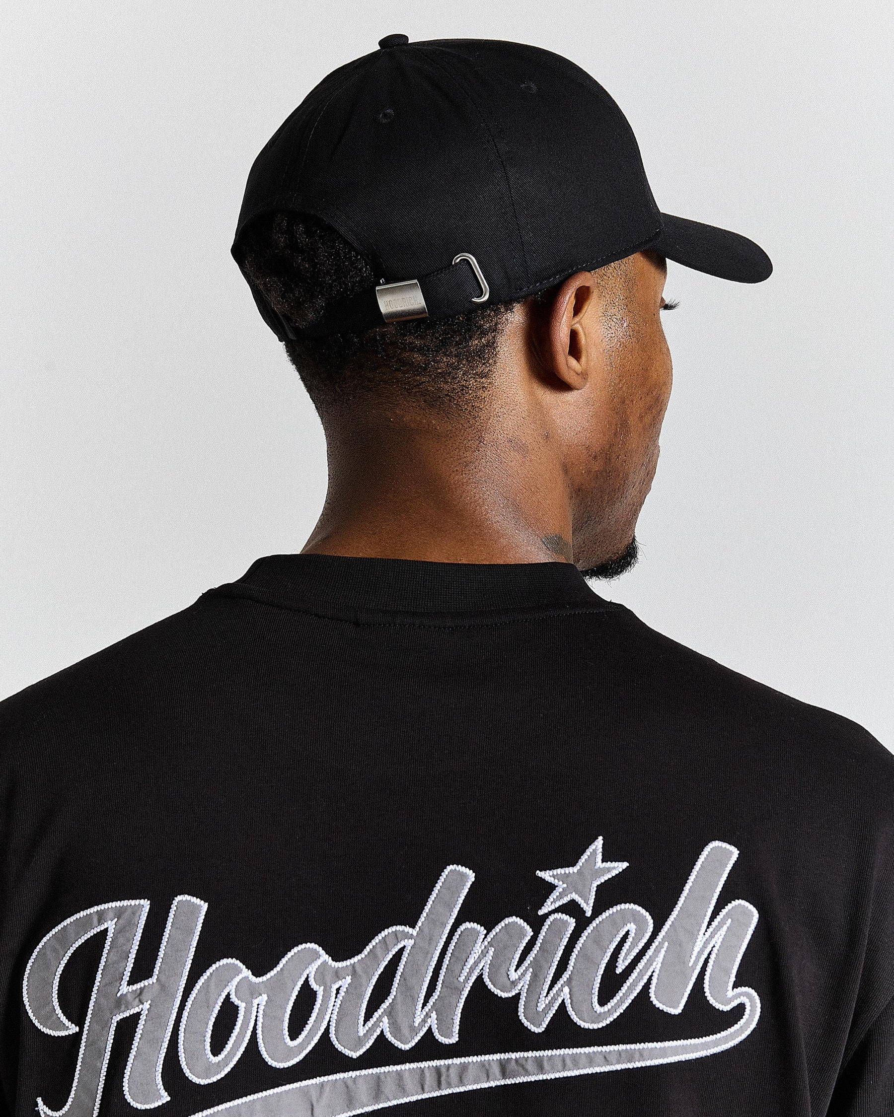 Person wearing a black cap and black 'Hoodrich' jacket on a light gray background