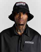 Person wearing a black 'Hoodrich' bucket hat and shirt against a gray background