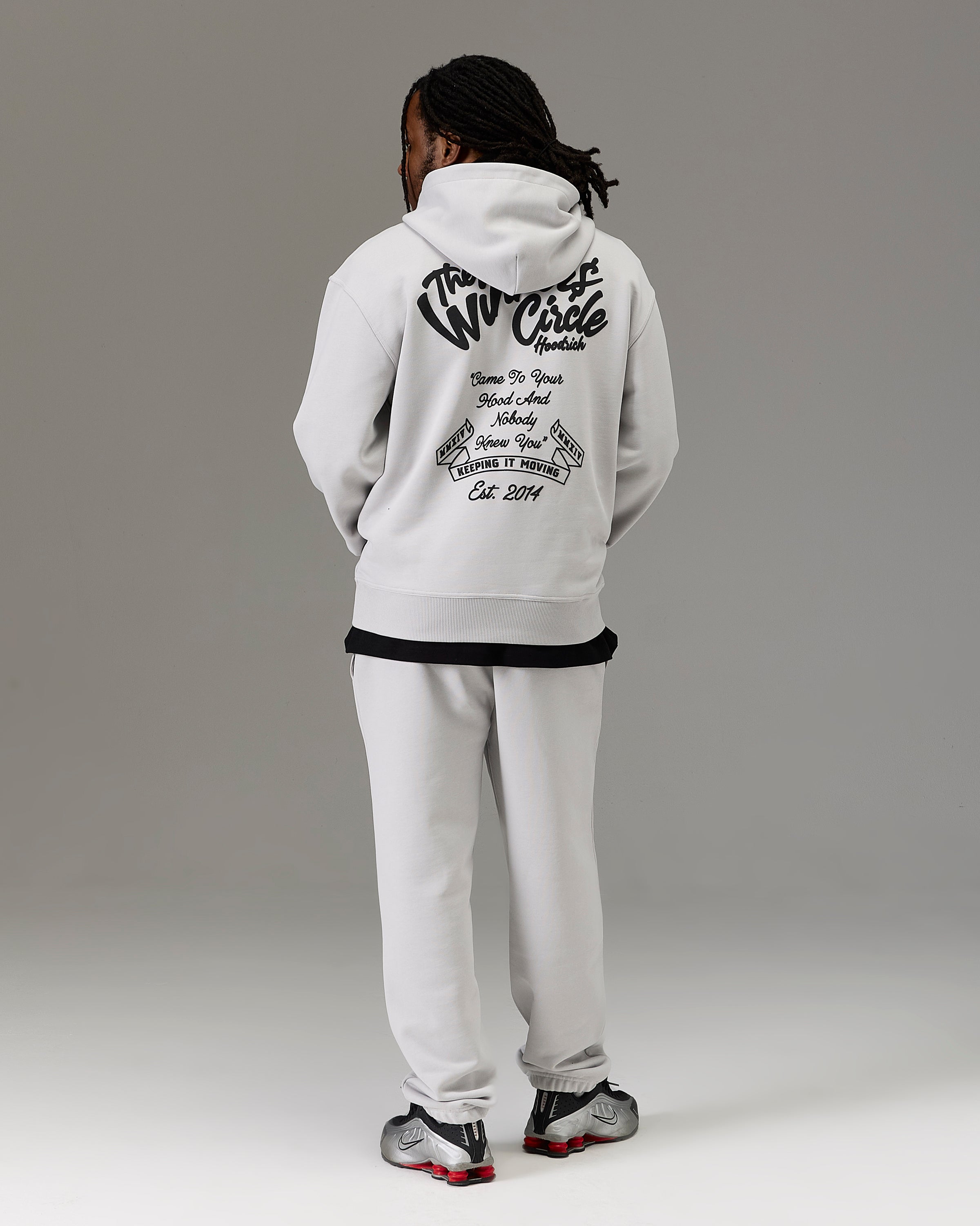 A model shown from the back wearing a white hoodie with a large black graphic print on the back, paired with white sweatpants and black and red sneakers, against a neutral gray studio background.