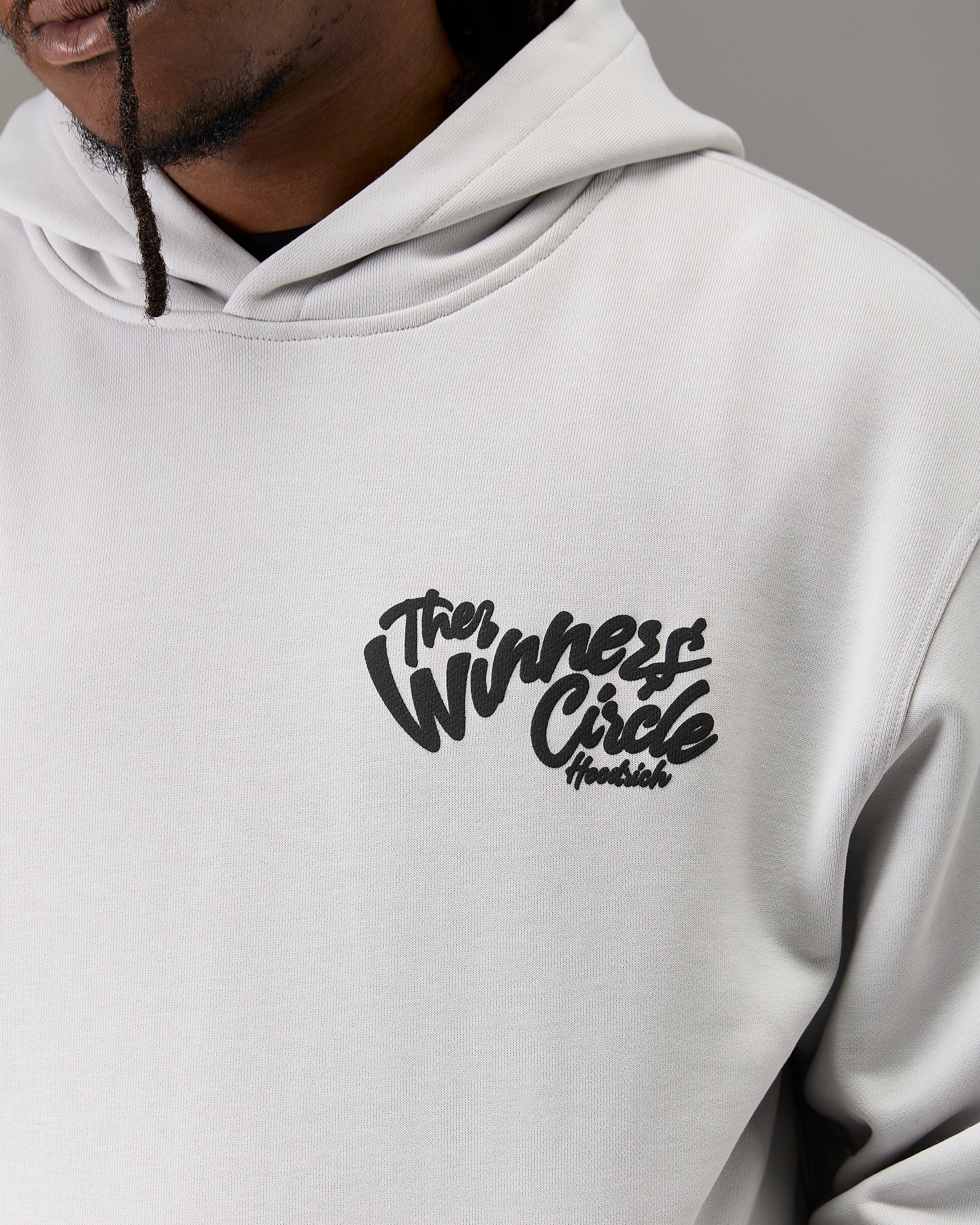 A close-up of a person wearing a light gray hoodie with a black graphic on the left chest that reads 'The Winners Circle Hoodies'.