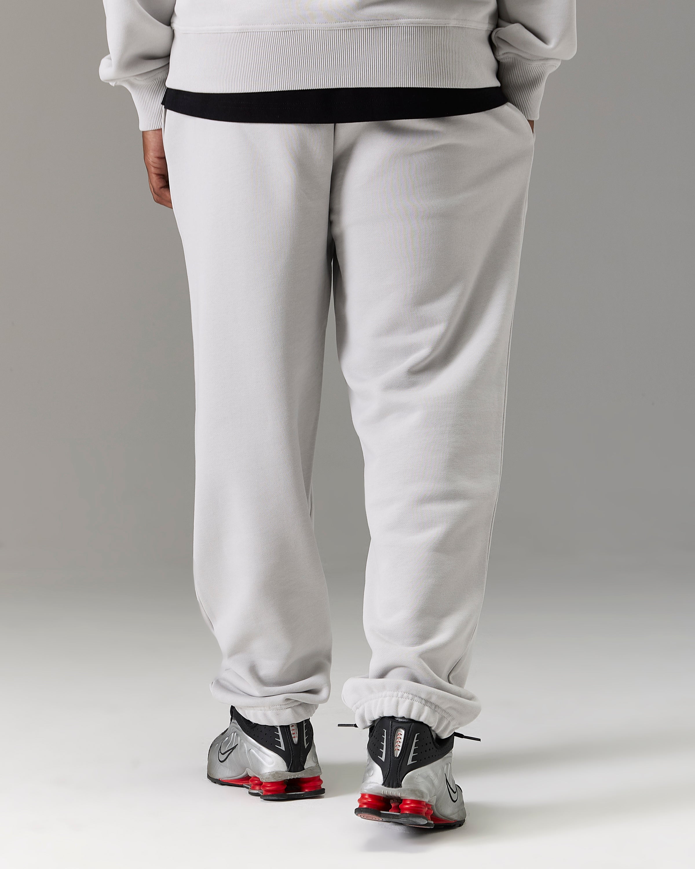 A person shown from the back wearing light gray sweatpants with a black waistband, a light gray sweatshirt, and gray sneakers with red accents, against a neutral gray studio background.