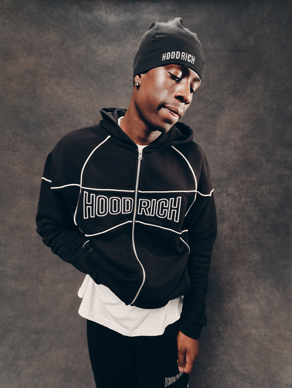 A man wearing a black zip-up hoodie with 'HOOD RICH' printed across the chest in white outlined letters, paired with a matching black beanie and black pants, standing against a textured dark gray studio background.