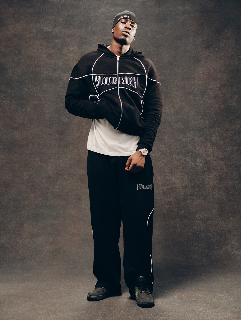 A man modeling a black zip-up hoodie with white graphic lines and 'HOODRICH' text across the chest, paired with black sweatpants, a white t-shirt, and a black beanie against a textured brown studio backdrop.