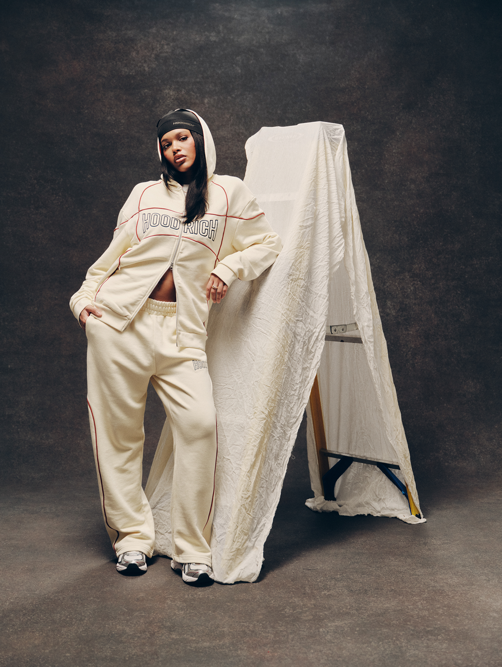 A model wearing a cream-colored HOODRICH tracksuit with red piping details stands next to a large white garment bag against a dark textured studio backdrop.