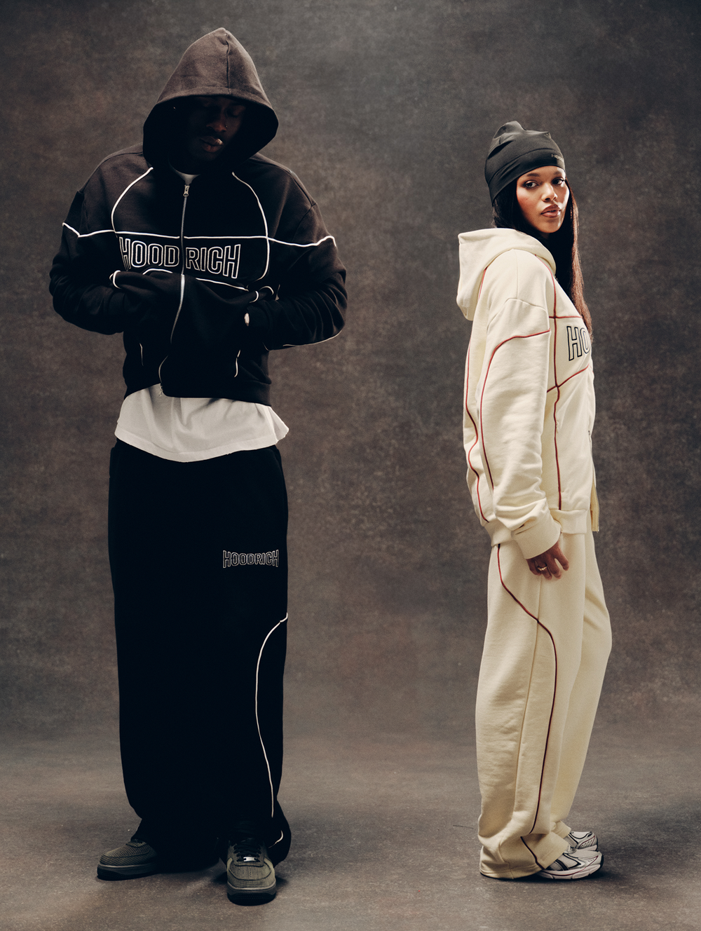 Two models posing in streetwear tracksuits from the brand HOODRICH. The model on the left wears a black zip-up hoodie with white piping and 'HOODRICH' text across the chest, paired with black sweatpants.