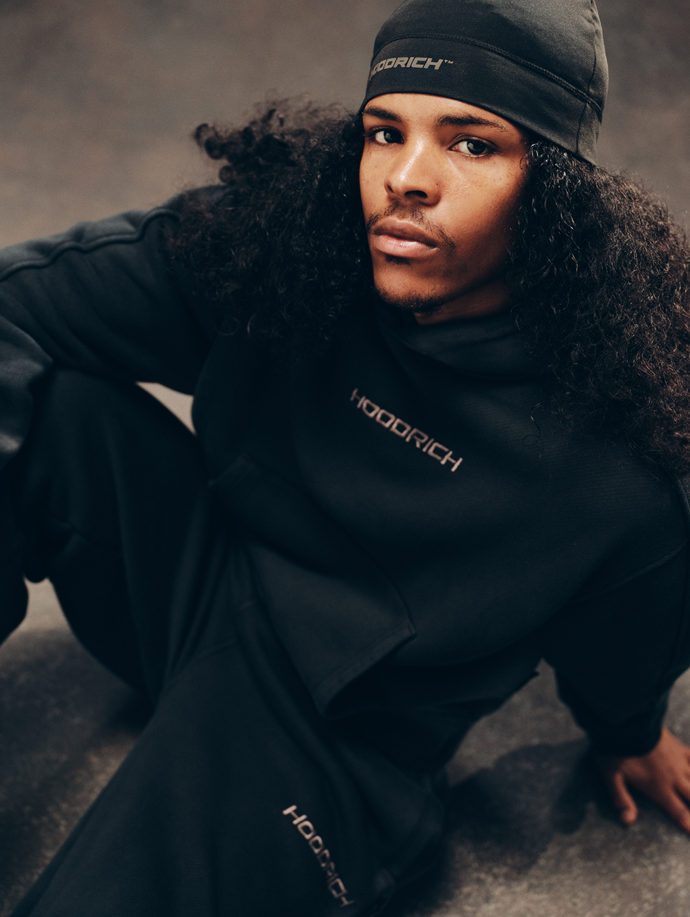 A model wearing a black hooded sweatshirt and matching pants with gold 'HOODRICH' branding, paired with a black beanie, posed on a textured concrete floor.