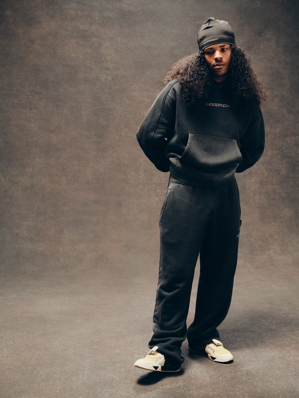 A model wearing a black hooded sweatshirt and matching sweatpants with white sneakers, standing against a textured brown studio backdrop.